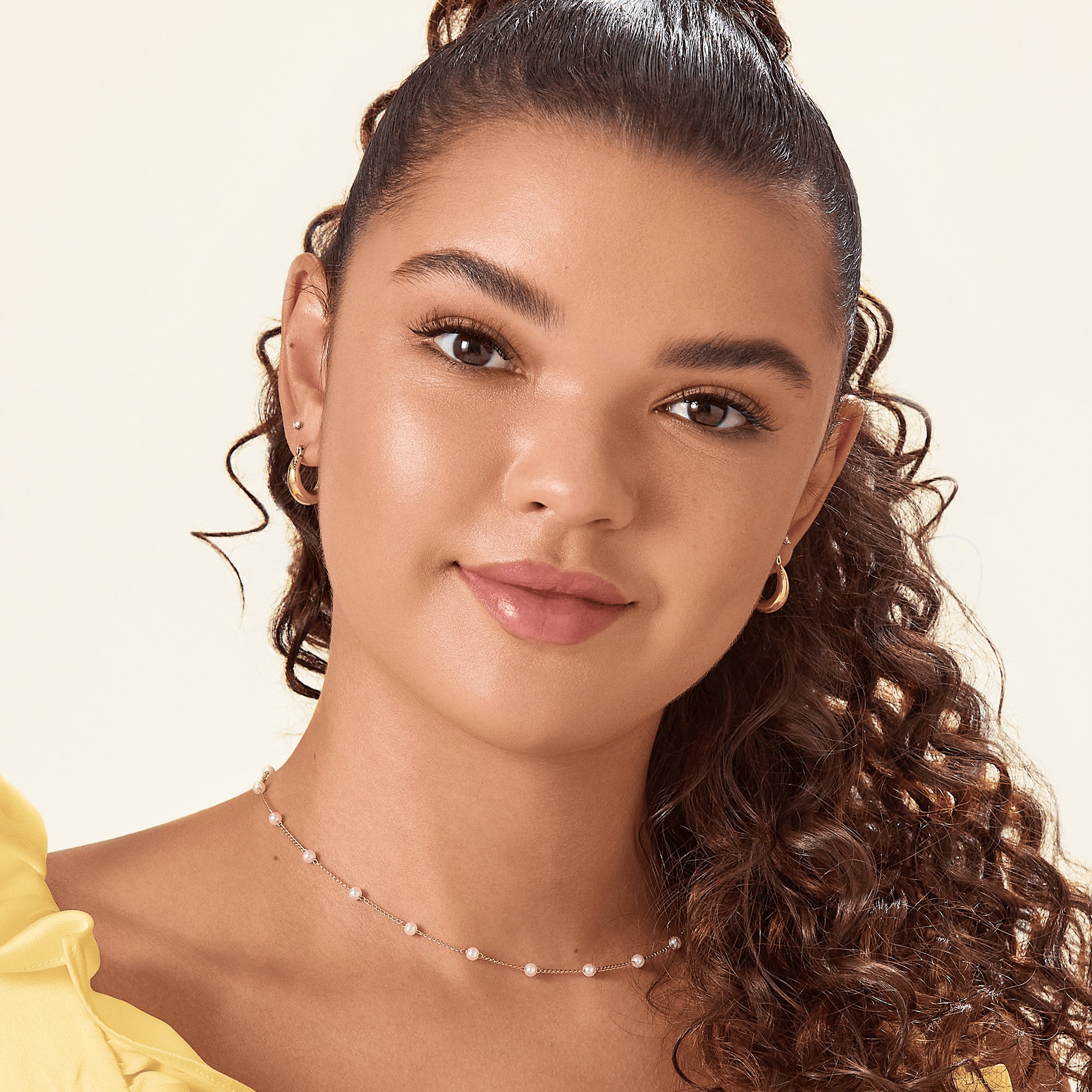 Close-up of a woman with warm-toned skin wearing natural-looking self-adhesive false eyelashes that enhance her upper lash line, styled with gold hoop earrings, a delicate pearl necklace, and a yellow ruffled top against a neutral background.
