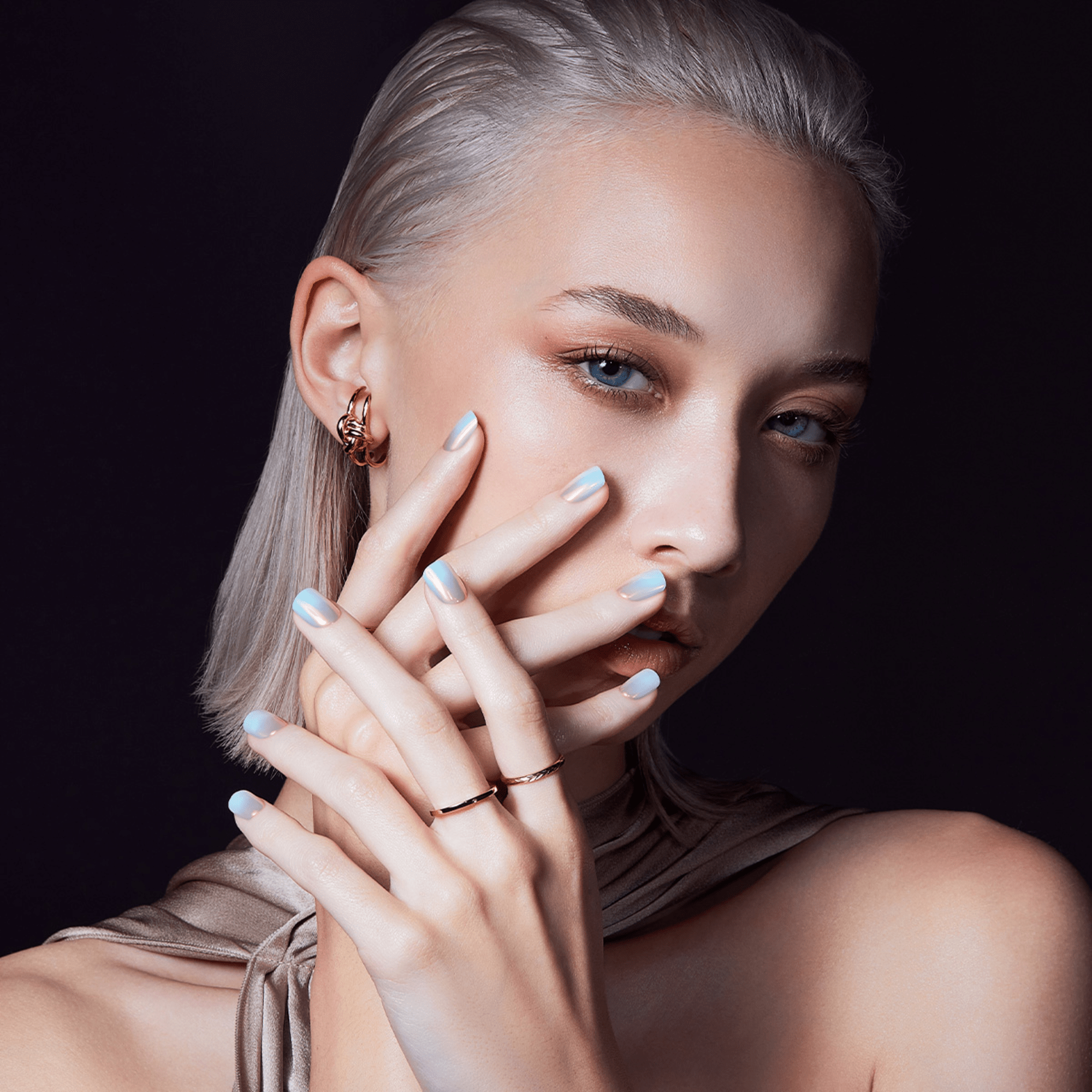 The image features a person with light blonde hair styled in a sleek, straight manner. They have a fair complexion and are posing with their hands near their face, showcasing a manicure with light-colored nail polish. The person is wearing a gold ring on one finger and a gold earring. The background is dark, which contrasts with the person&