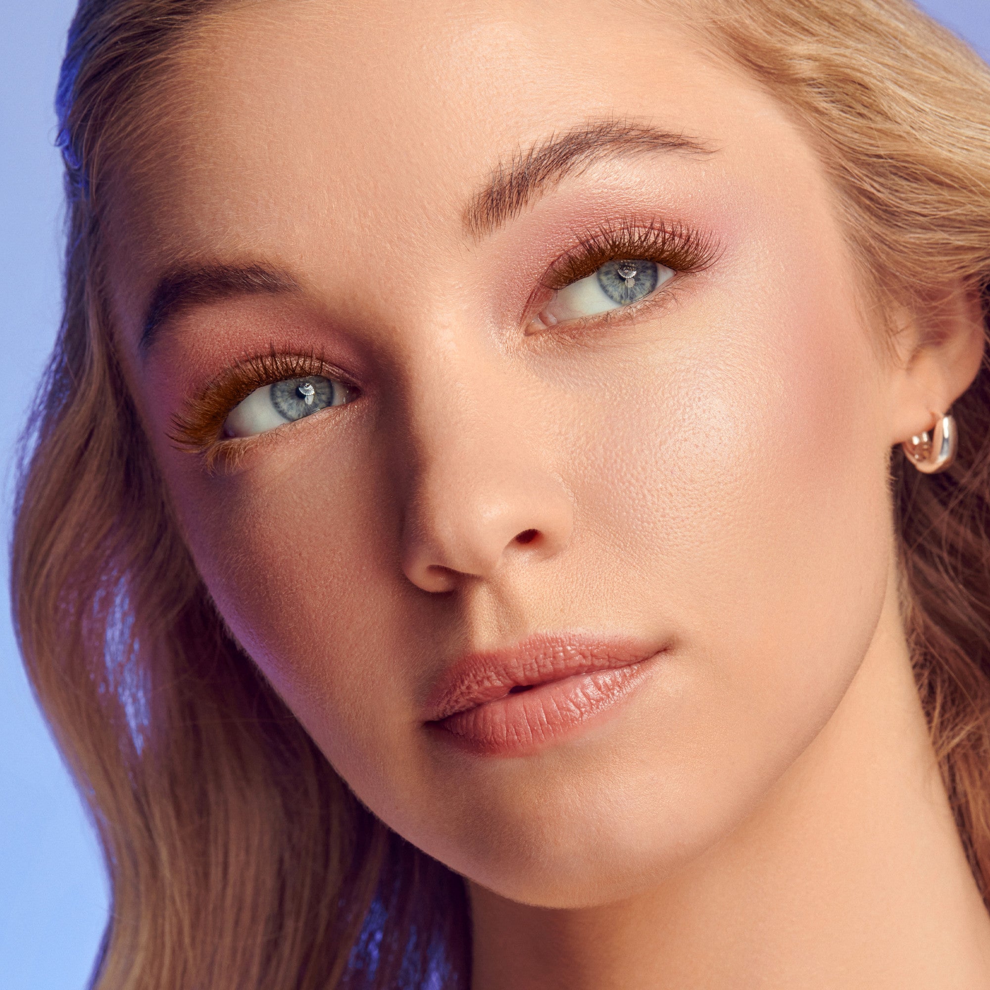Close-up of a woman with fair skin and blue eyes wearing chestnut brown false eyelash wisps on her upper and lower lashes, with soft pink eyeshadow and nude lipstick.