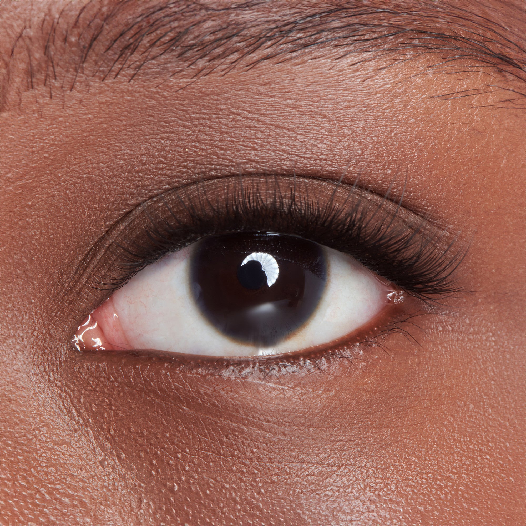 The image features a close-up of a human eye. The eye has a dark brown iris and a prominent pupil, surrounded by long, dark eyelashes. The skin around the eye is smooth and has a warm, brown tone. The overall focus is on the eye, highlighting its details and textures.