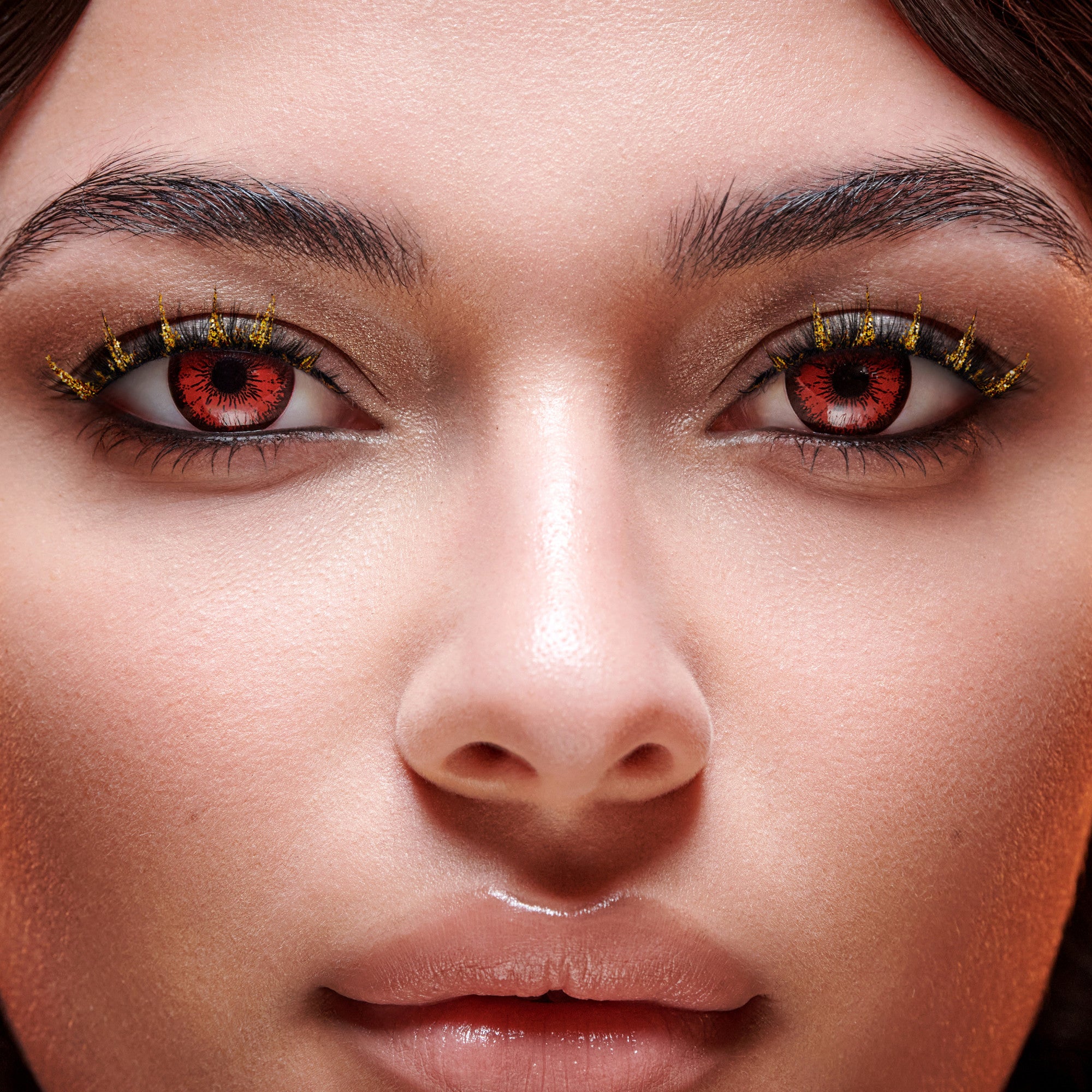 Close-up of a person’s face showcasing Halloween-themed decorative lashes with gold glitter accents and red contact lenses with black radial designs.