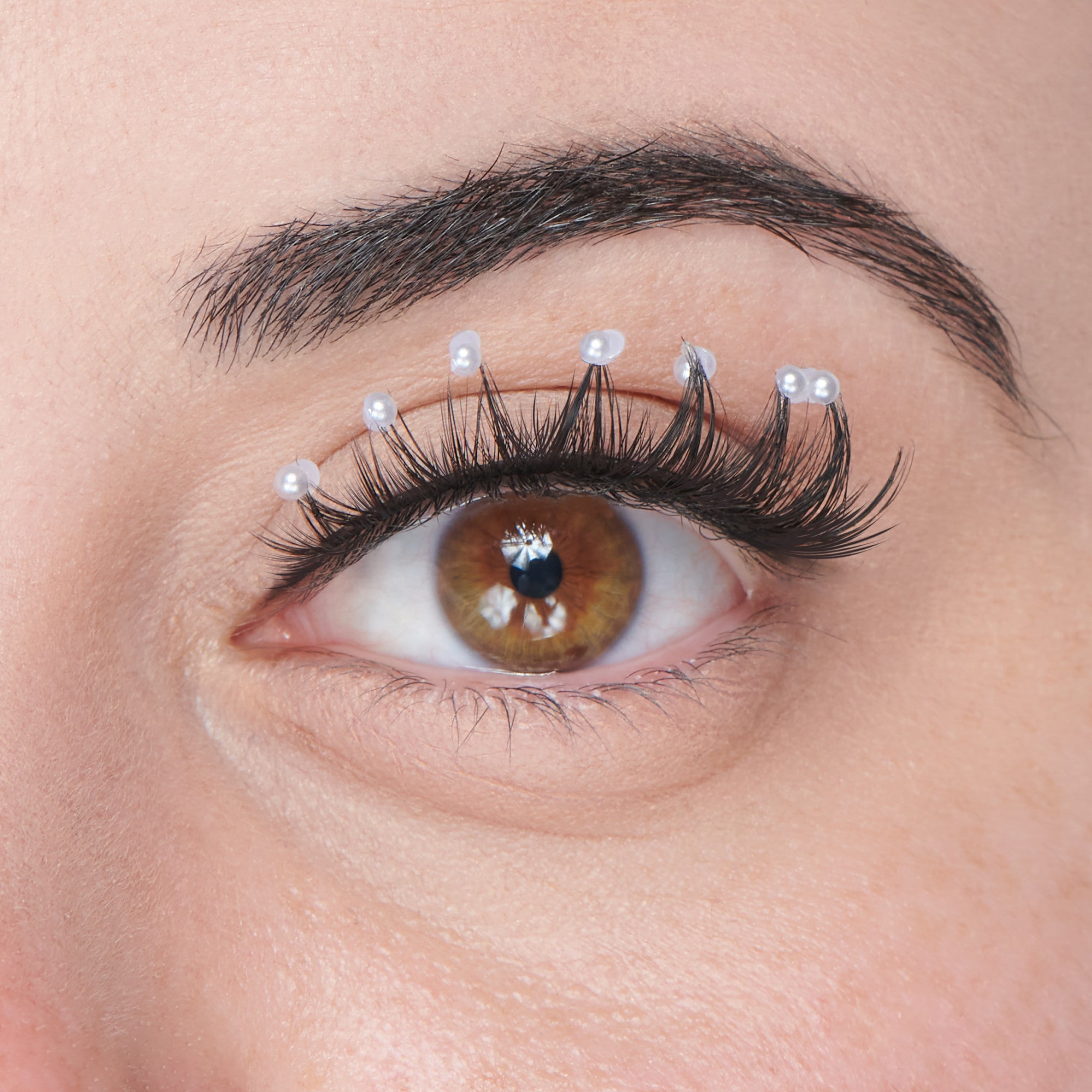 Close-up of a brown eye with long, black, curled false eyelashes decorated with small white pearl-like gem stickers along the upper lash line.