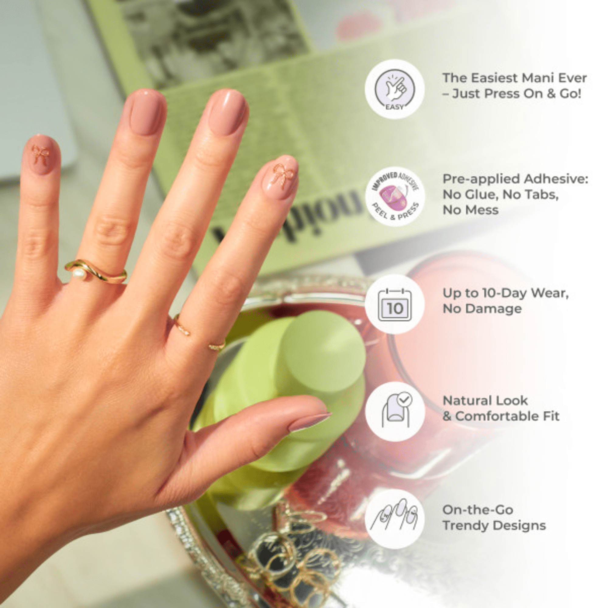 Hand with natural pink press-on nails featuring small gold bow designs on the pinky and ring finger, shown over a tabletop with skincare items.