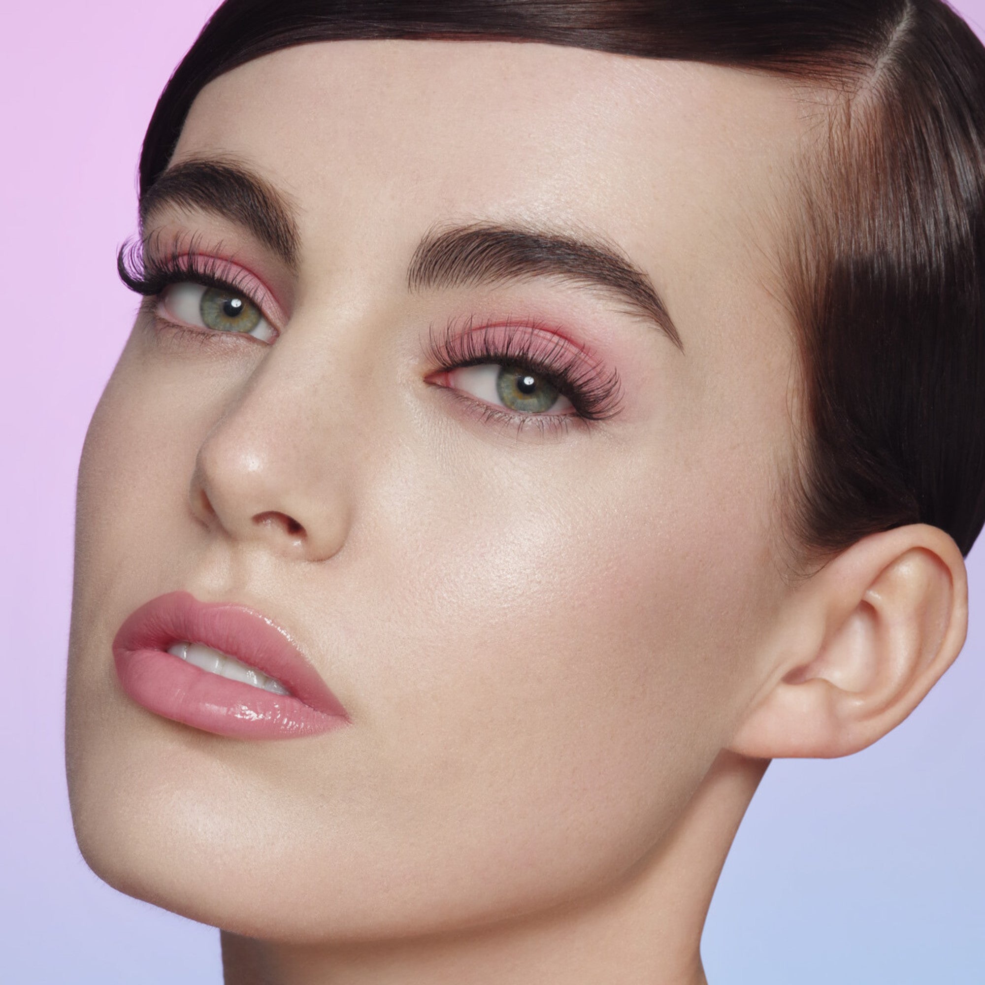 Close-up of a woman’s face showing long, wispy 18mm false eyelashes with pink eyeshadow and glossy pink lips, set against a soft purple background.