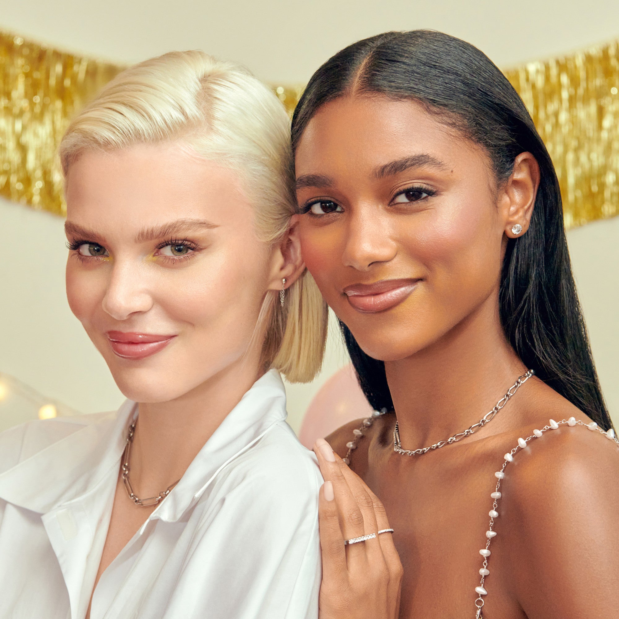 The image features two women standing closely together, both smiling. One woman has short, platinum blonde hair and is wearing a white shirt, while the other has long, straight black hair and is dressed in a delicate, embellished outfit. They are adorned with elegant jewelry, including earrings and necklaces. The background includes festive decorations, such as a gold garland and soft lighting, creating a cheerful atmosphere.