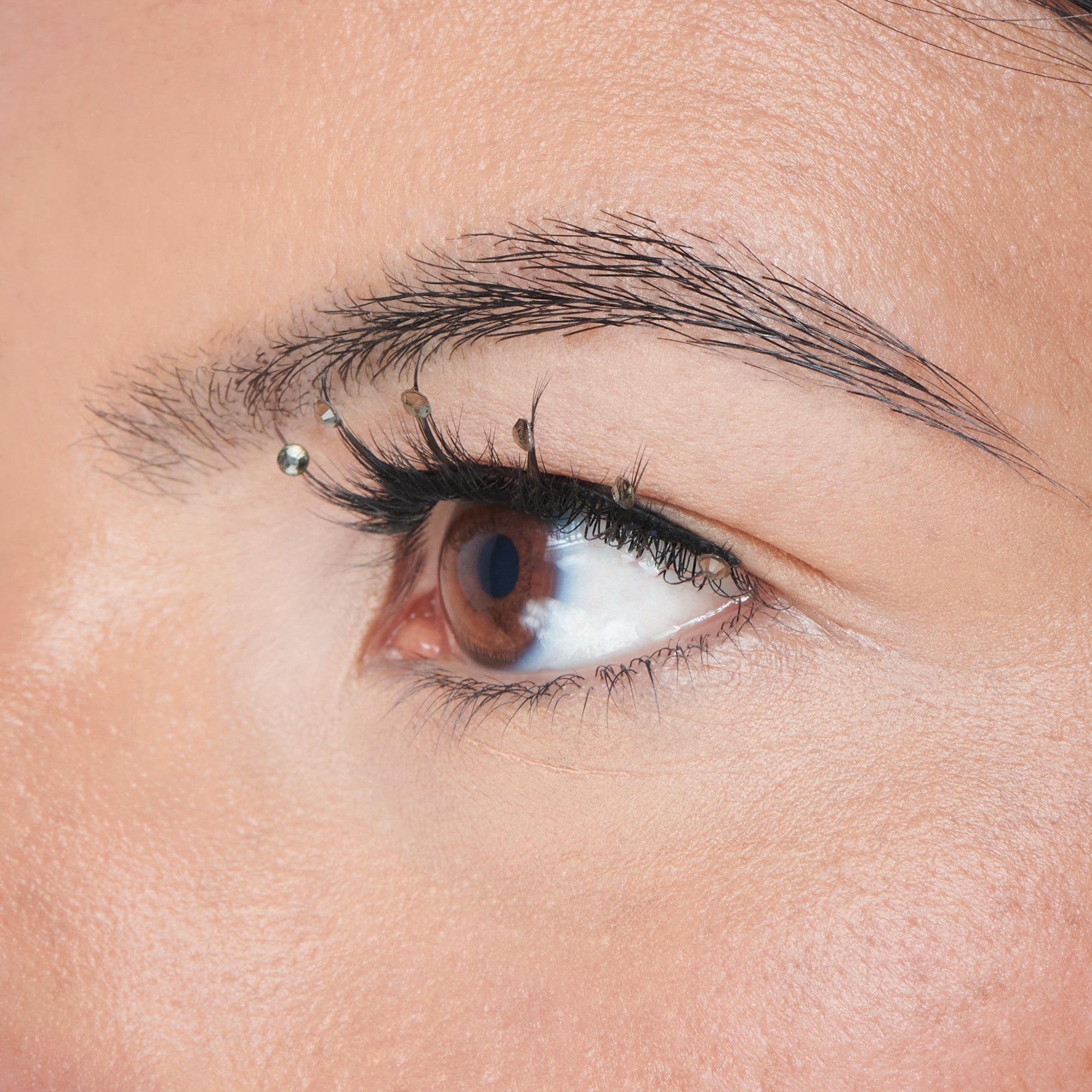 Close-up side view of a brown eye with long black decorated lashes adorned with small clear gem stickers along the upper lash line.