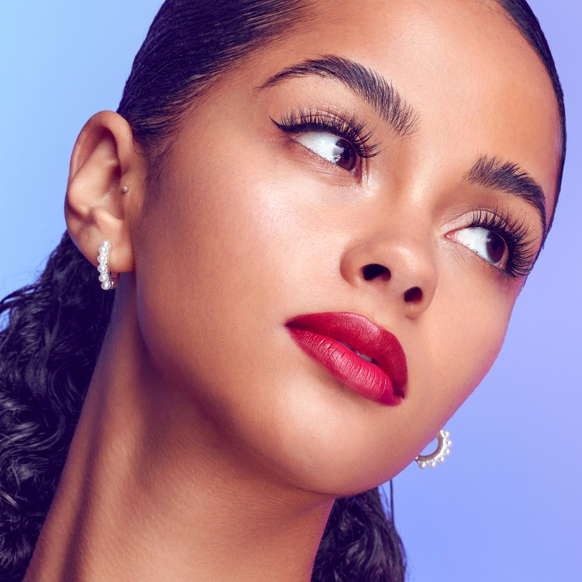 Close-up of a woman with smooth skin and bold red lipstick wearing voluminous, long lash extensions against a soft purple background.