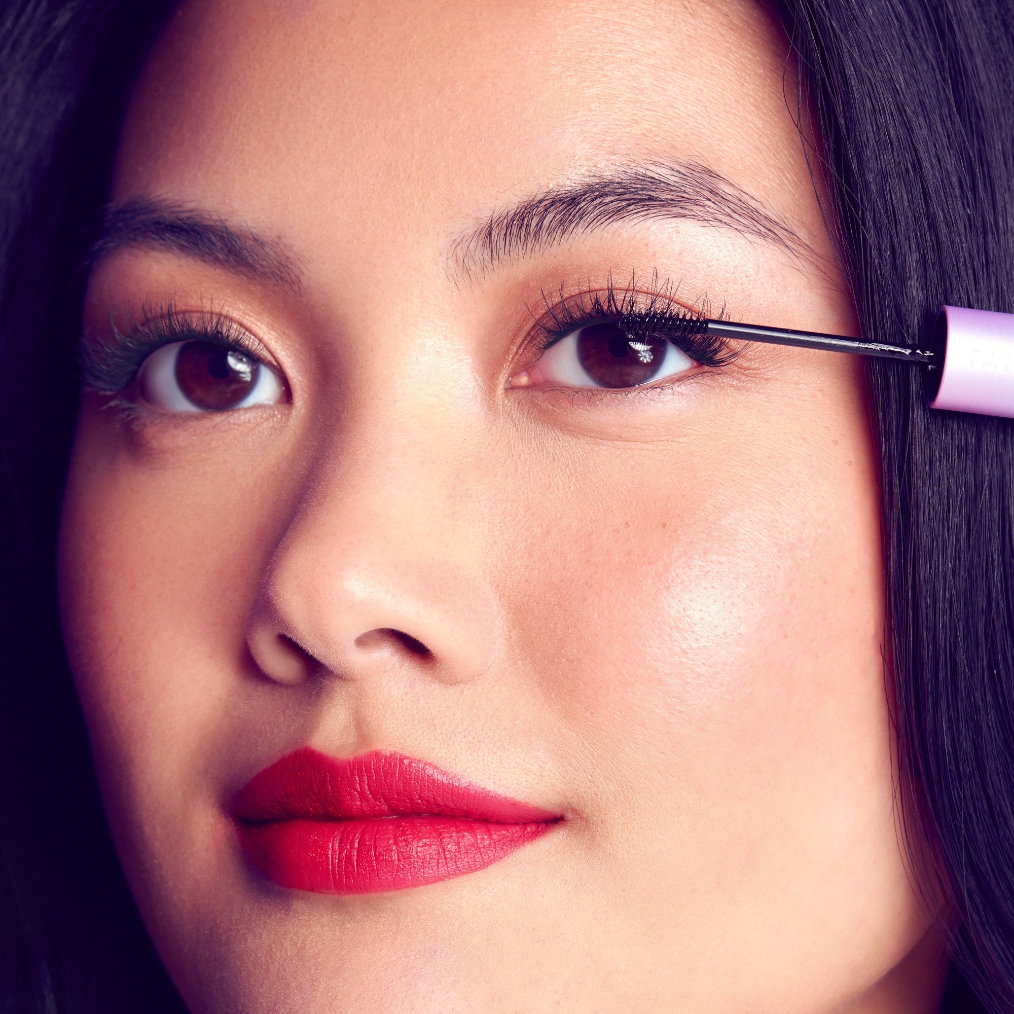 Close-up of a woman applying FALSCARA mascara to her upper eyelashes, showing long, defined lashes and bright red lipstick on smooth skin.