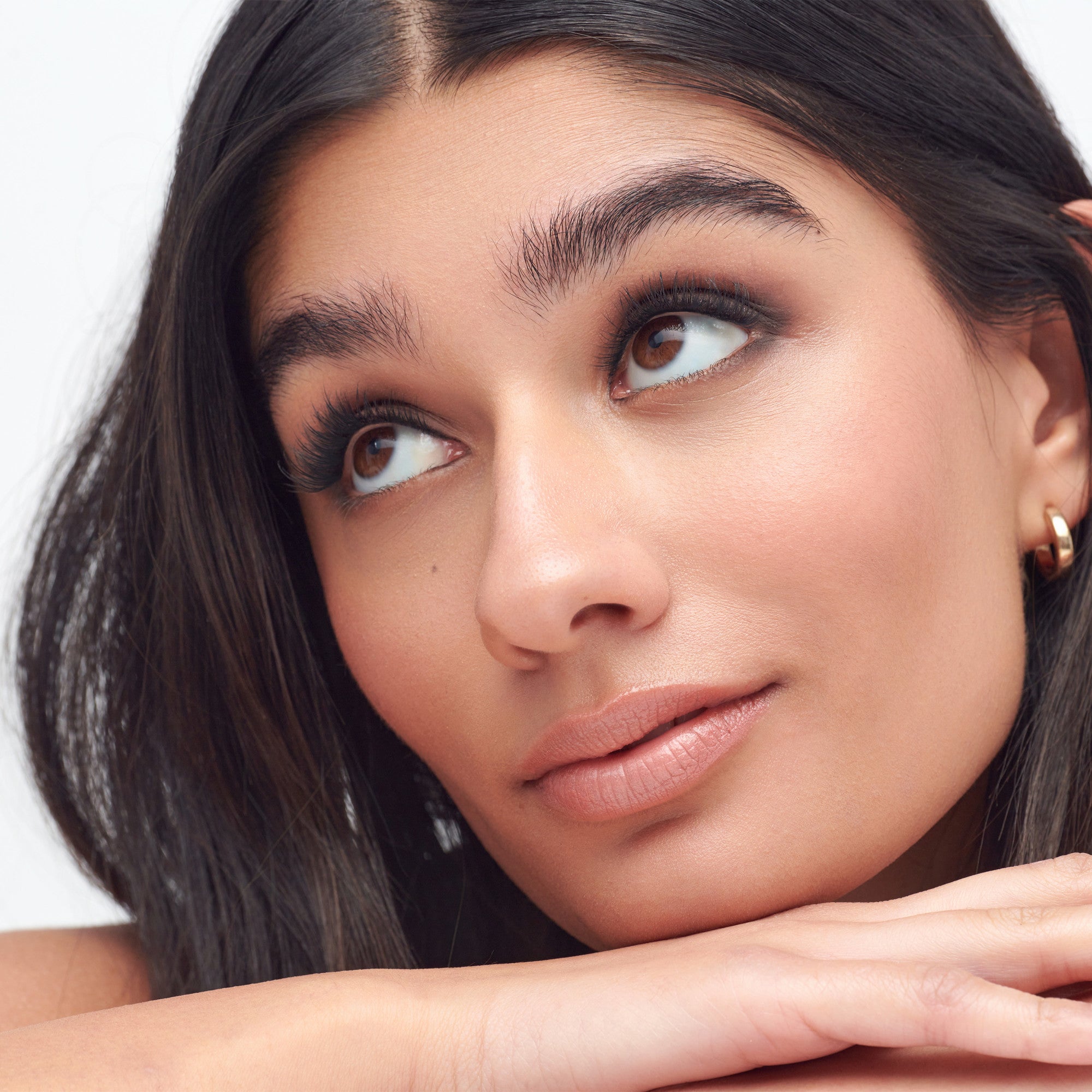 Close-up of a woman with medium skin tone and dark hair resting her chin on her hands, showcasing long, thick, curled false eyelashes and natural makeup.