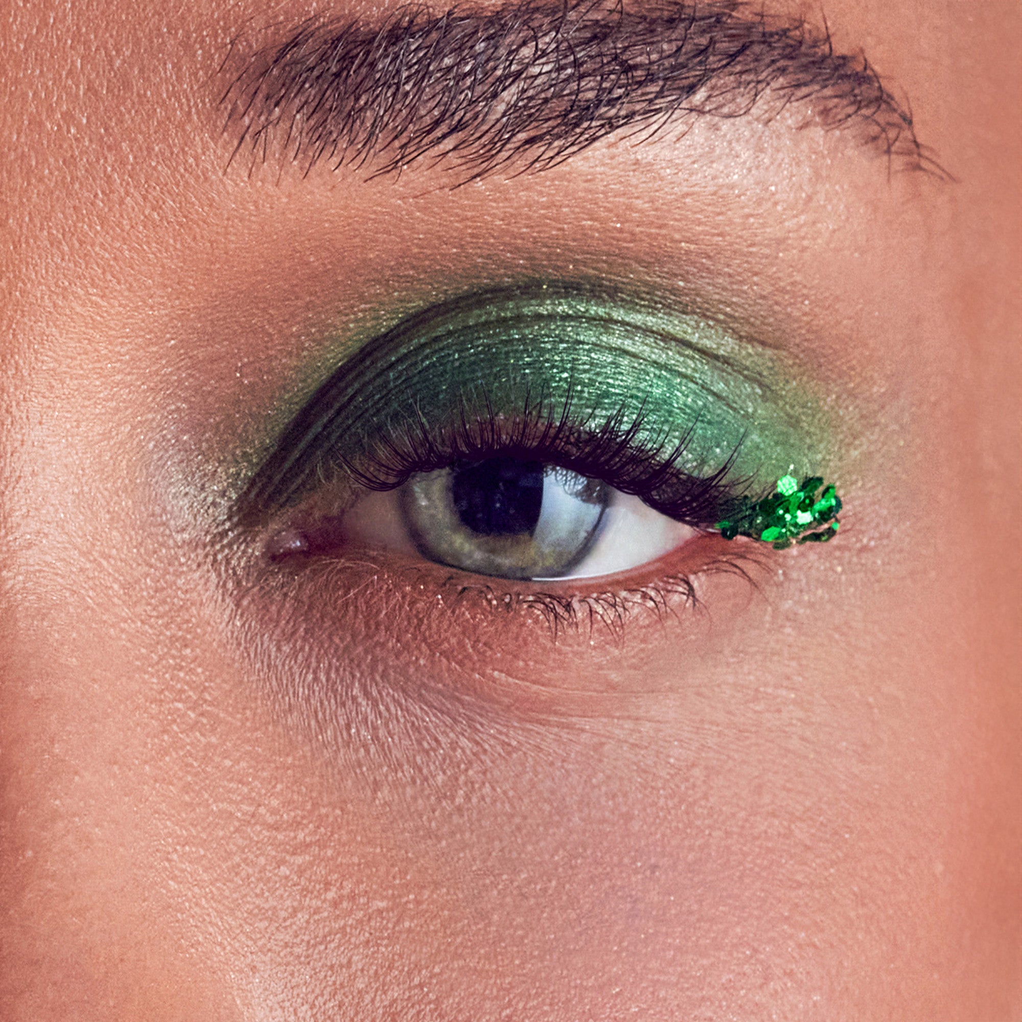 Close-up of an eye with long, dark false eyelashes and shimmering green eyeshadow extending from the eyelid to the outer corner, which is decorated with small green sequins.