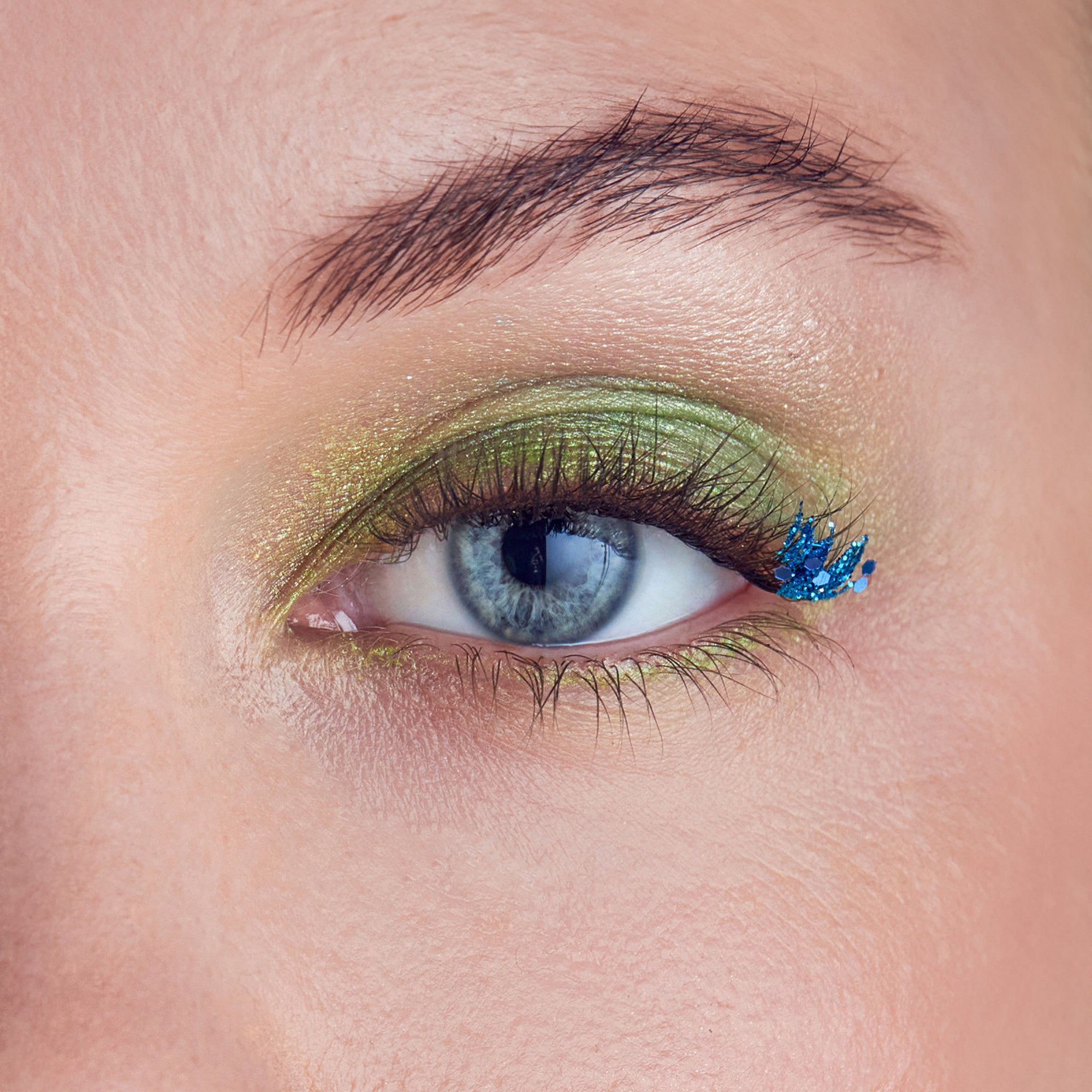 Close-up of a blue eye with green metallic eyeshadow and a unique false eyelash style featuring clustered blue glitter tips on the outer corner.