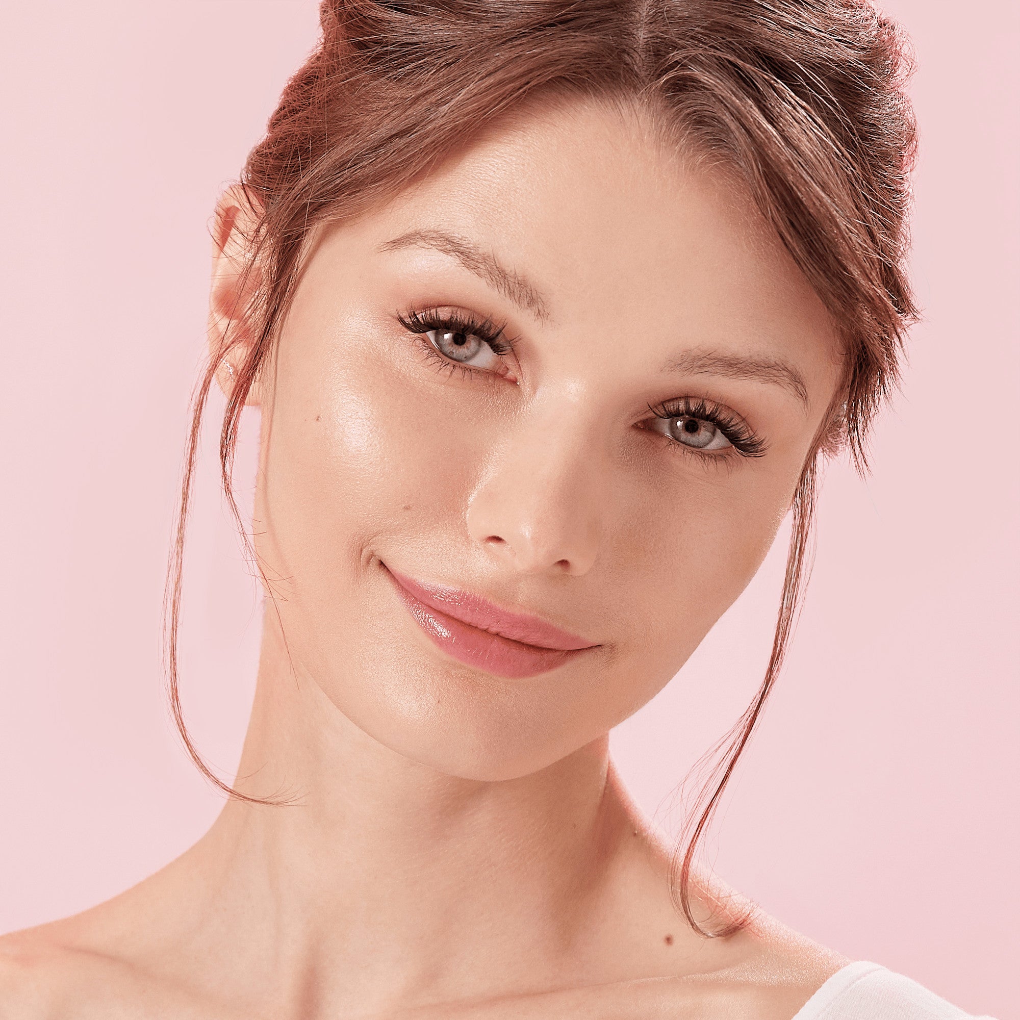 Close-up of a young woman with fair skin and light brown hair wearing bold, dark, self-adhesive false eyelashes, smiling against a soft pink background.