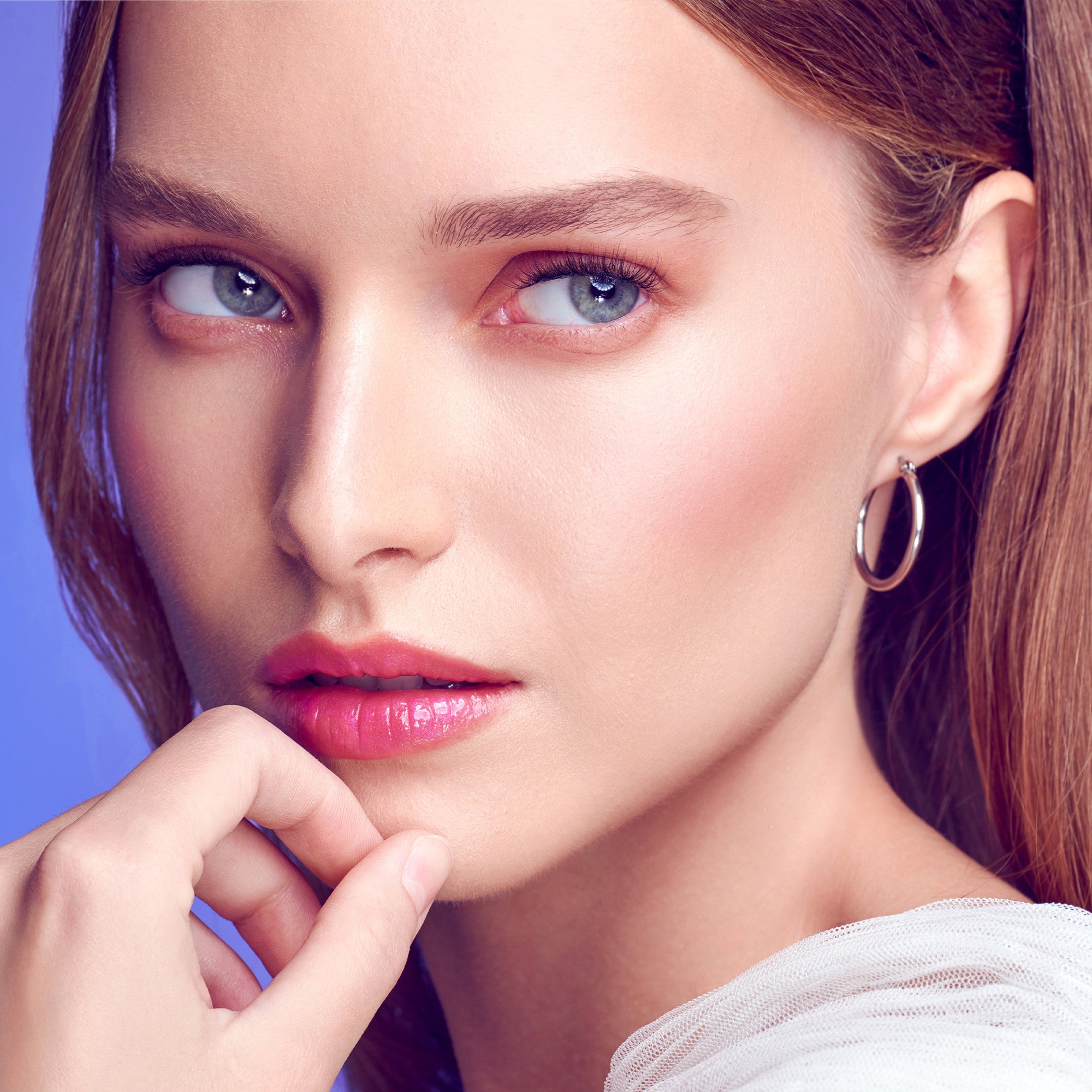 Close-up of a woman with fair skin, light blue eyes, and natural-looking lash extensions, wearing silver hoop earrings and a white textured top.