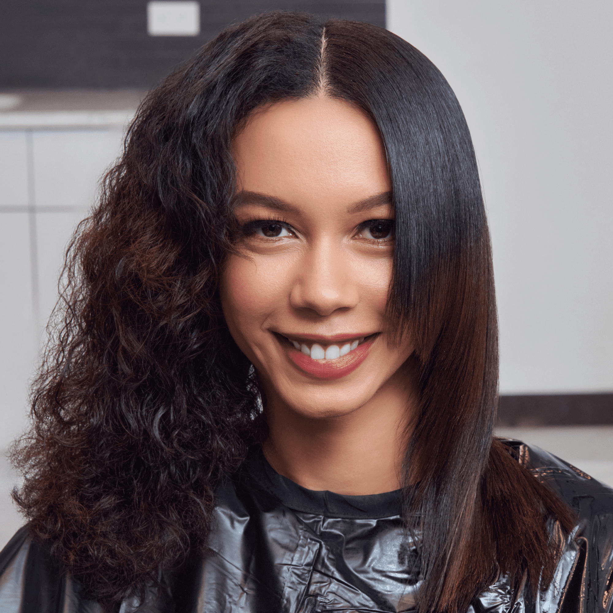 Smiling woman with half curly, half smooth straightened dark hair, wearing a black salon cape, demonstrating hair transformation effect.