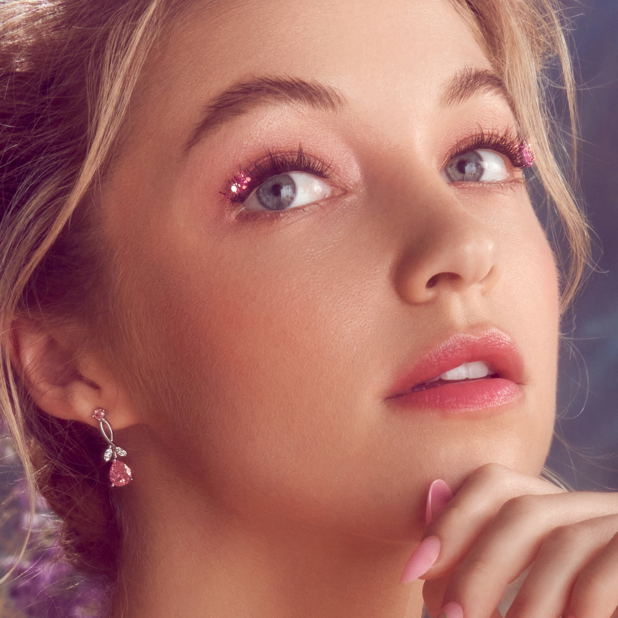 Close-up of a woman with fair skin and blue eyes wearing pink glittery false eyelashes on the outer corners, soft pink eyeshadow, glossy pink lips, and matching pink teardrop earrings. Her hand with long, pink manicured nails gently touches her chin.