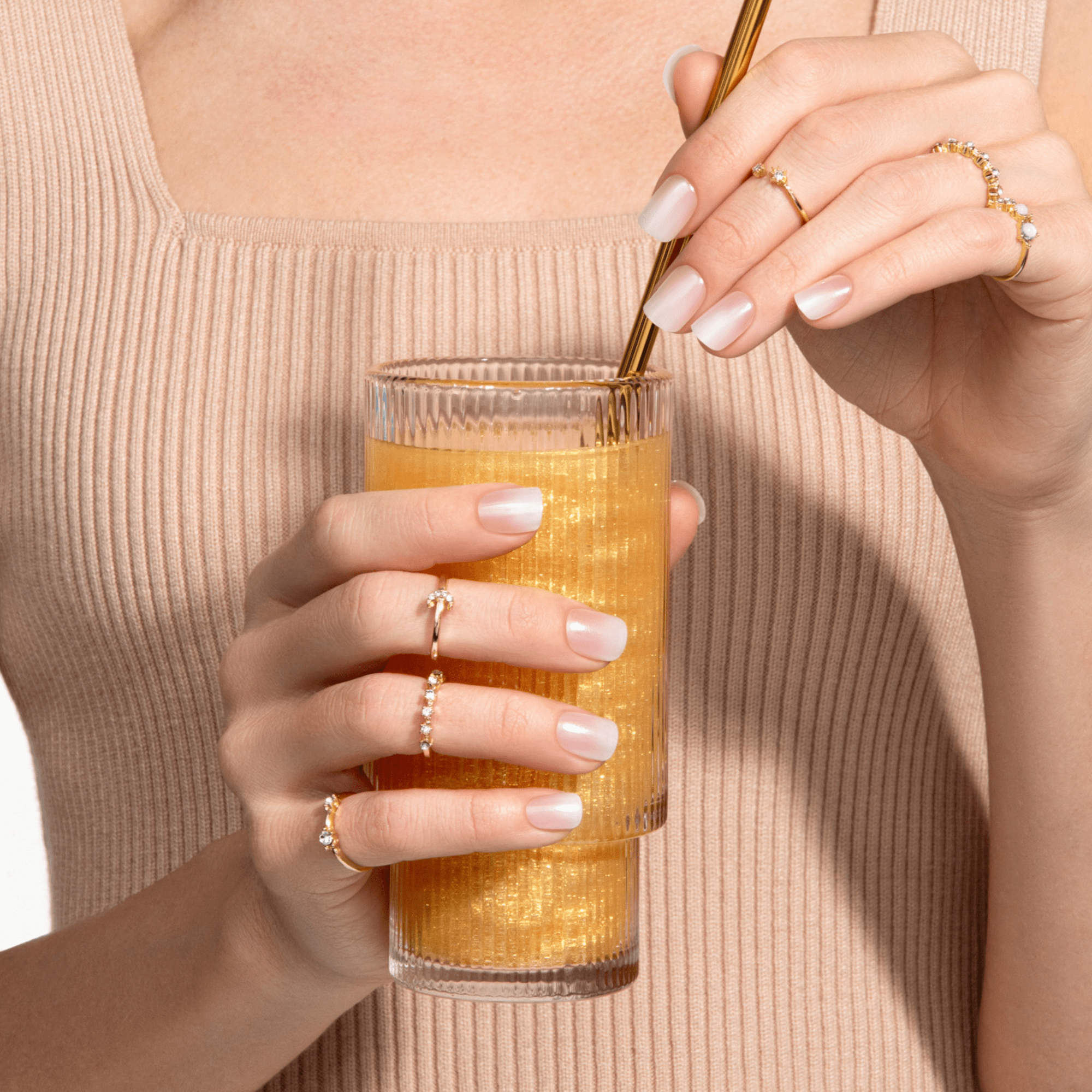 Hands with glossy, soft nude press-on nails holding a textured glass of orange-yellow beverage with a gold straw, against a beige ribbed top background.