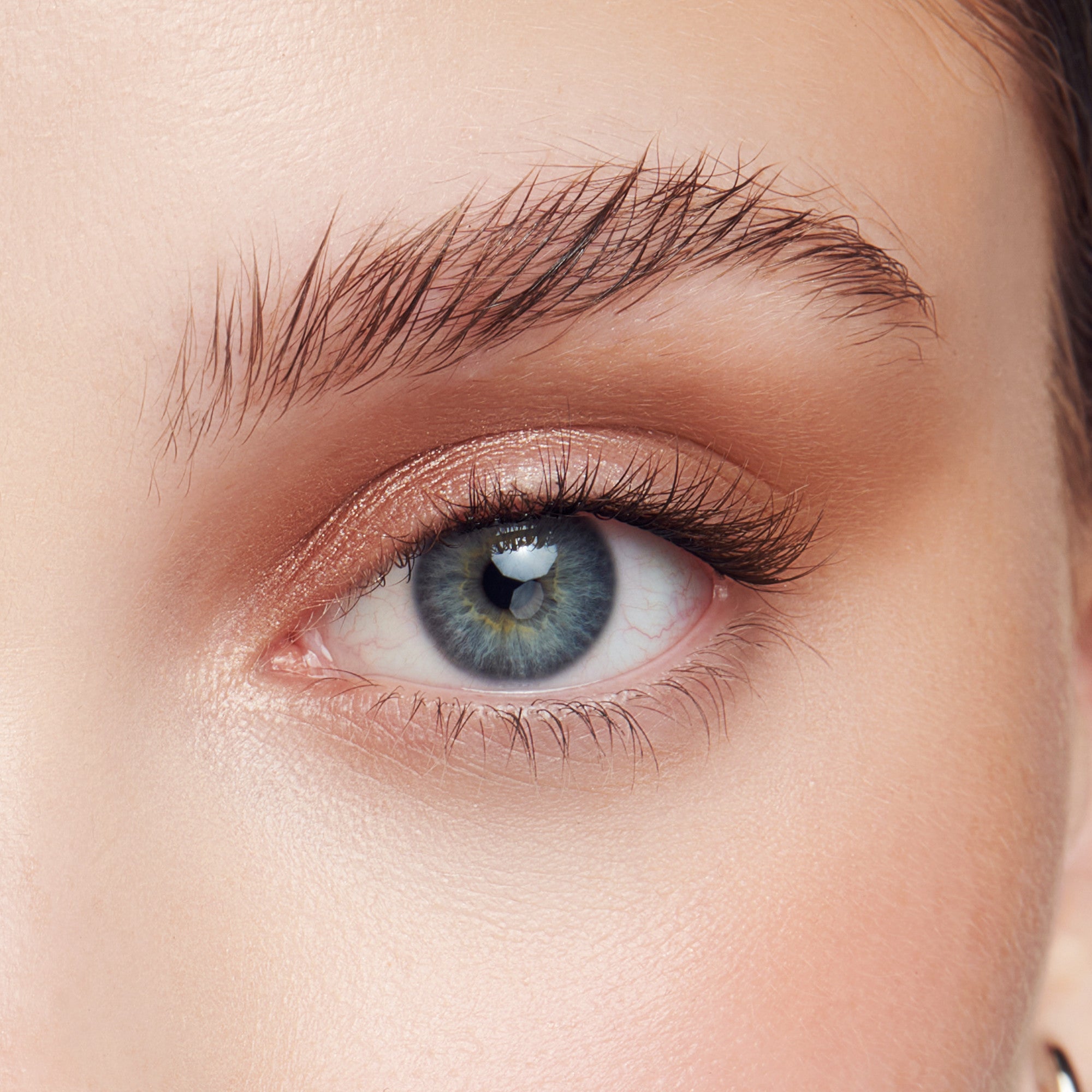 Close-up of a blue eye with airy, wispy false eyelashes applied, featuring natural-looking length and separation against soft, peach-toned eyeshadow.