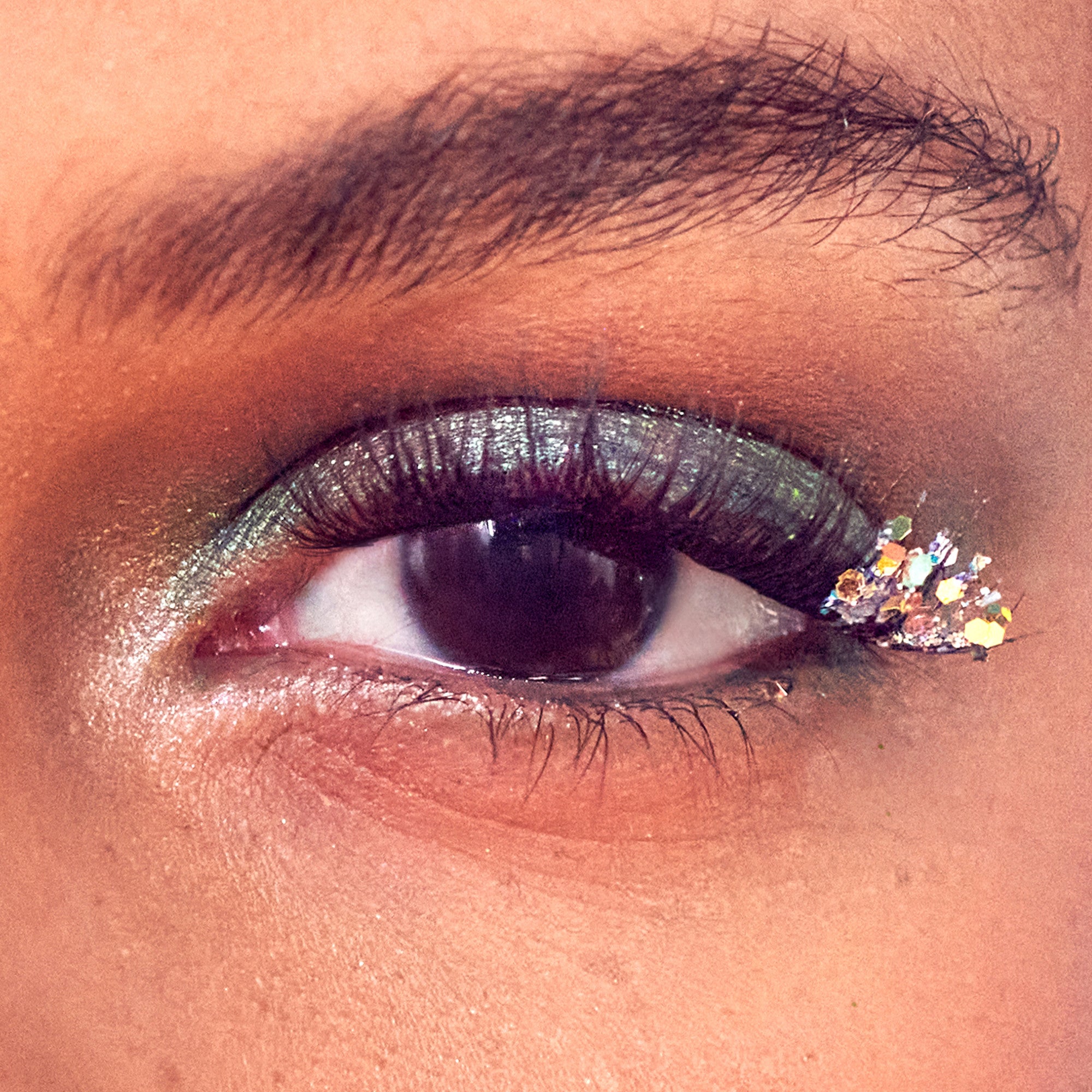 Close-up of a brown eye with shimmery green and silver eyeshadow and iridescent multi-shaped glitter accents at the outer corner, creating a sparkling, festive look.