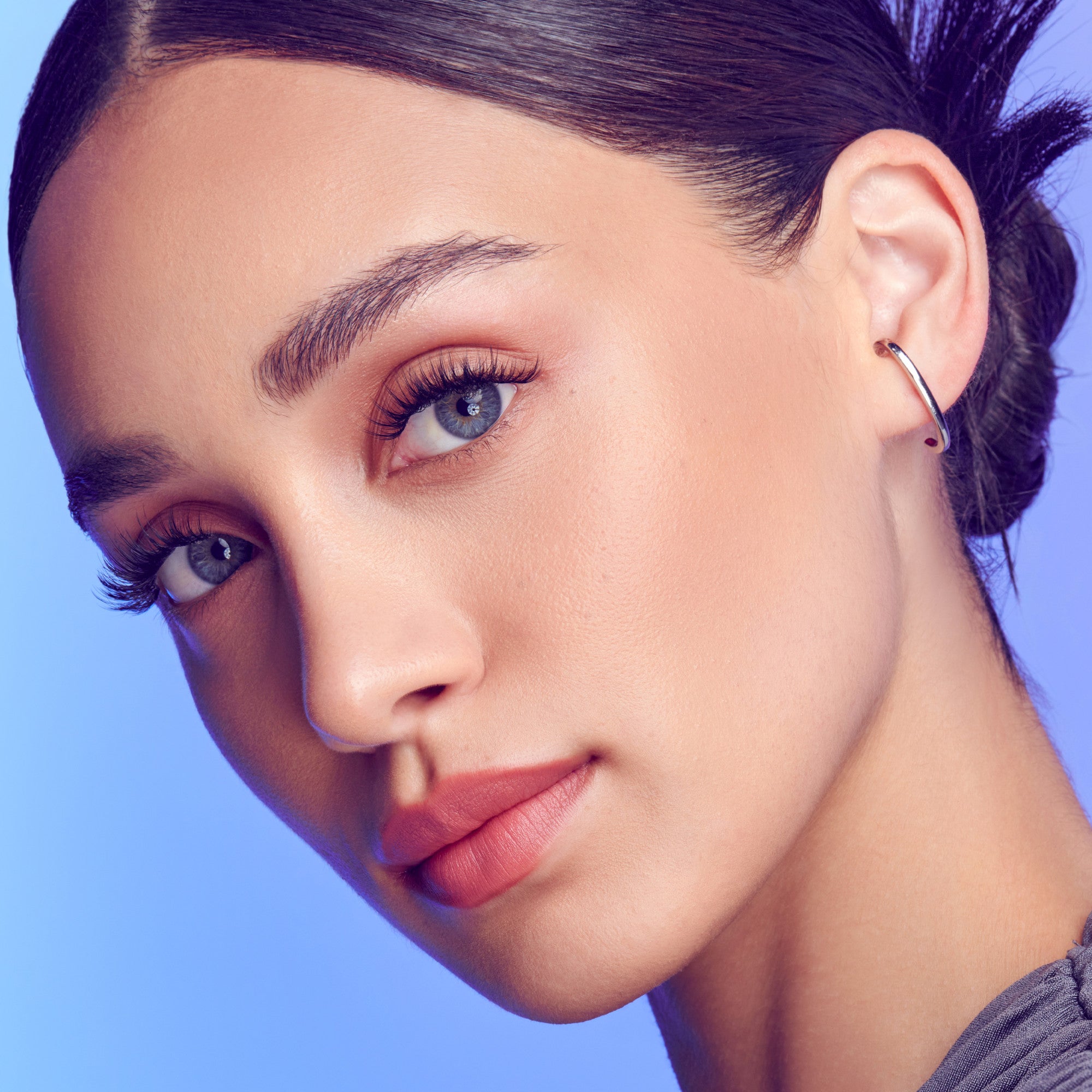 Close-up of a woman’s face showing delicate, natural-looking lash extensions with curled, wispy lashes against a soft blue background.