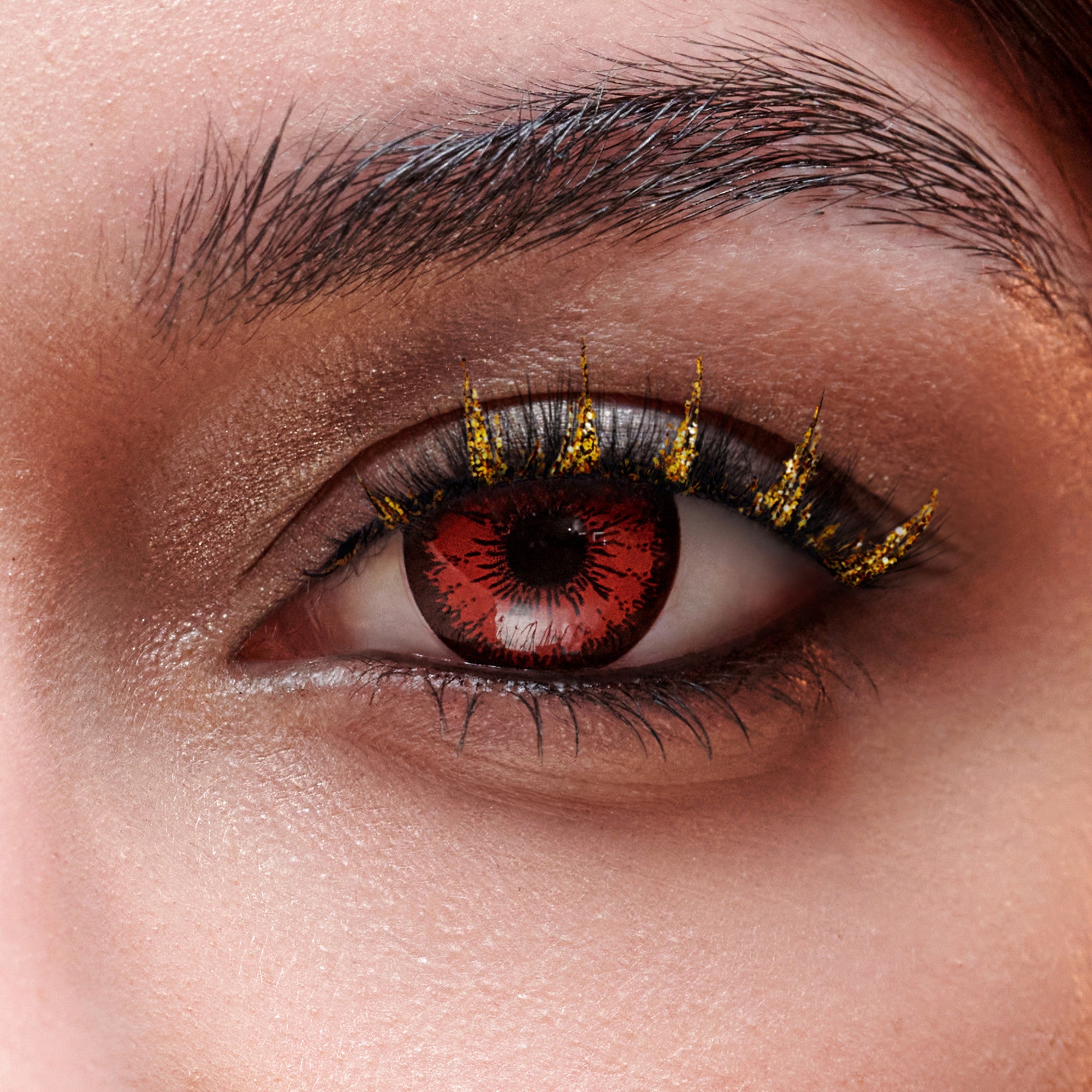 Close-up view of a human eye wearing red patterned Halloween contact lens and gold glitter decorated false lashes creating a dramatic, festive look.