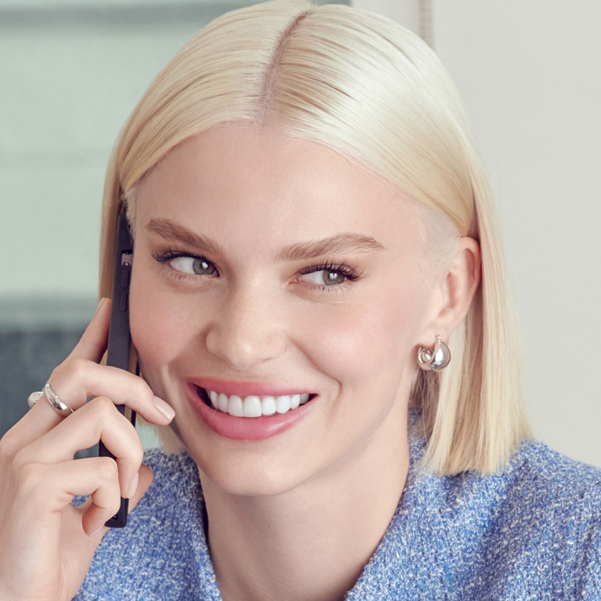 Close-up of a smiling woman with straight blonde hair wearing silver earrings and a blue textured top, showing natural-looking long eyelashes.