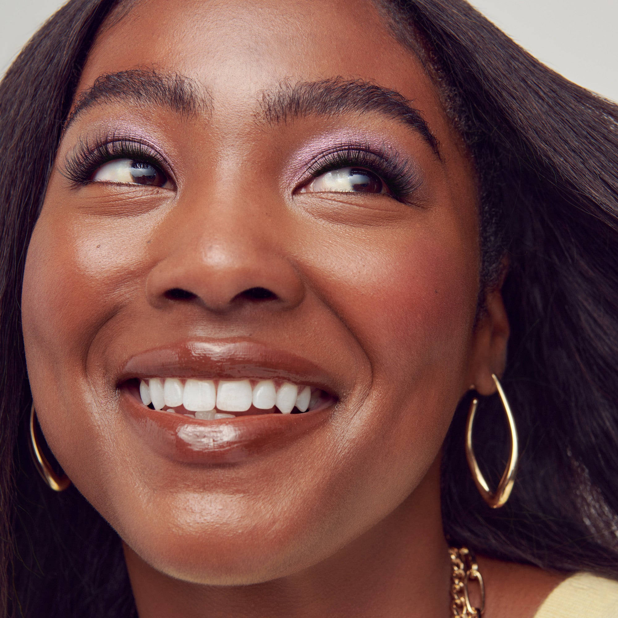 Close-up of a smiling woman with dark skin wearing wispy false eyelashes, soft purple eyeshadow, gold hoop earrings, and a gold chain necklace.