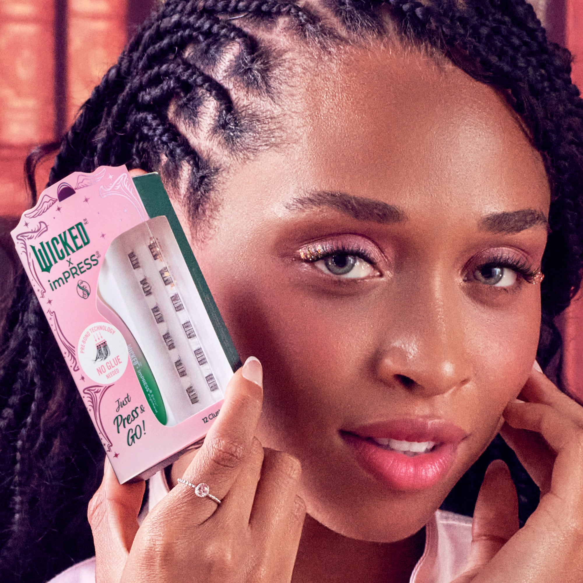 Woman with braided hair holds pink and green WICKED X imPRESS press-on lashes packaging near her face, highlighting \