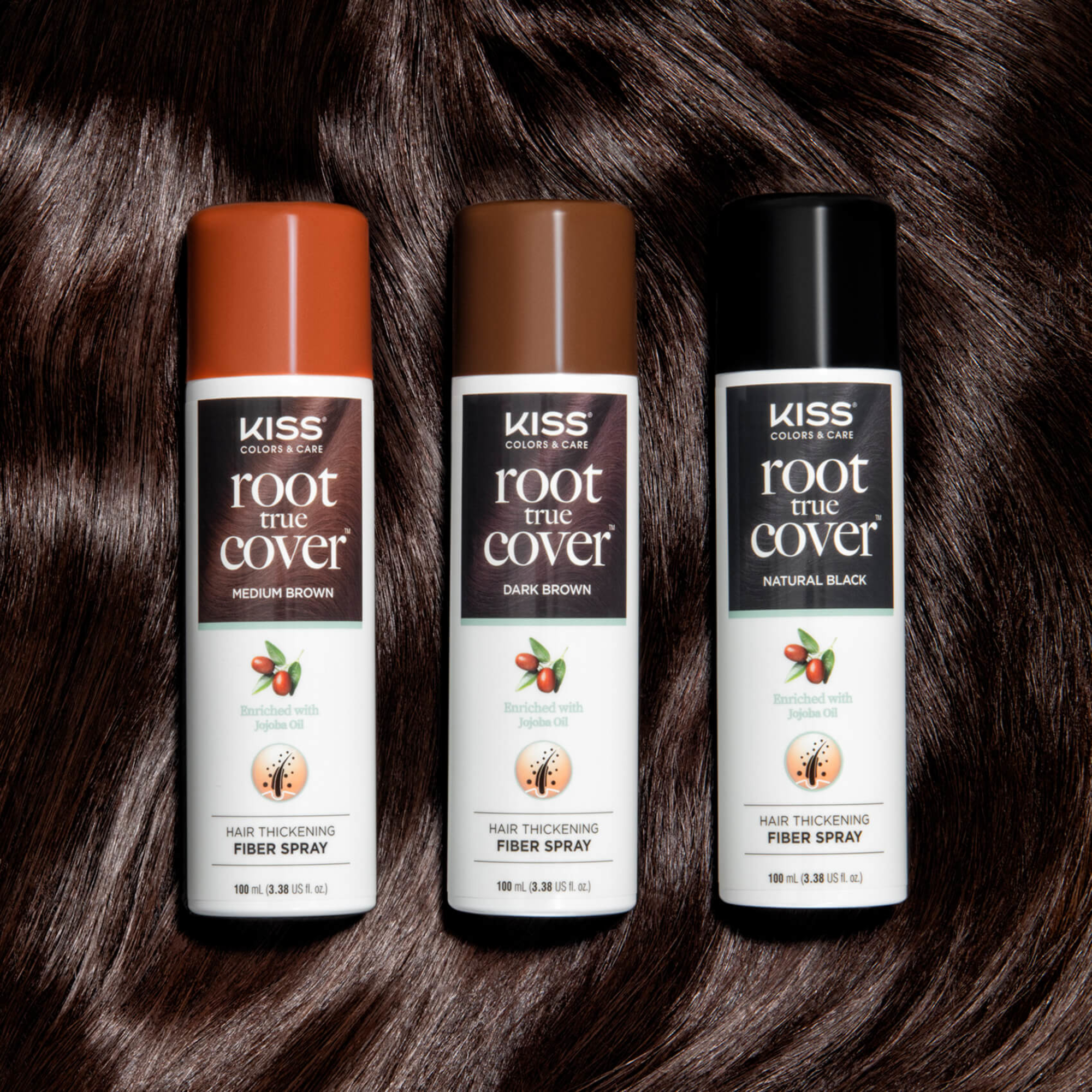Three KISS Colors & Care Root True Cover hair thickening fiber spray cans in Medium Brown, Dark Brown, and Natural Black, on a dark brown hair background, each labeled as enriched with jojoba oil.