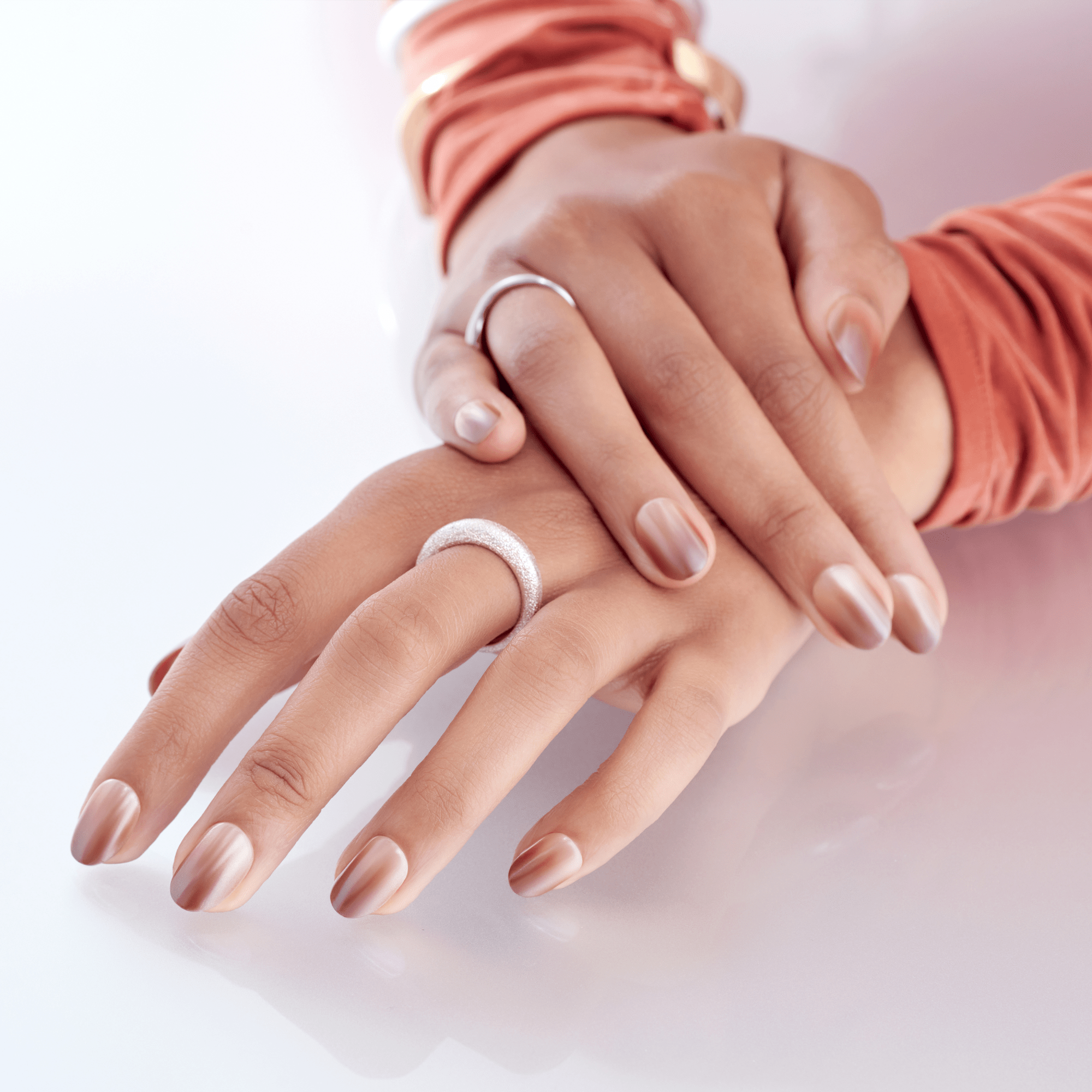 Hands with medium skin tone wearing press-on nails in a glossy gradient rose gold shade, displayed against a white surface with peach sleeves and silver rings.