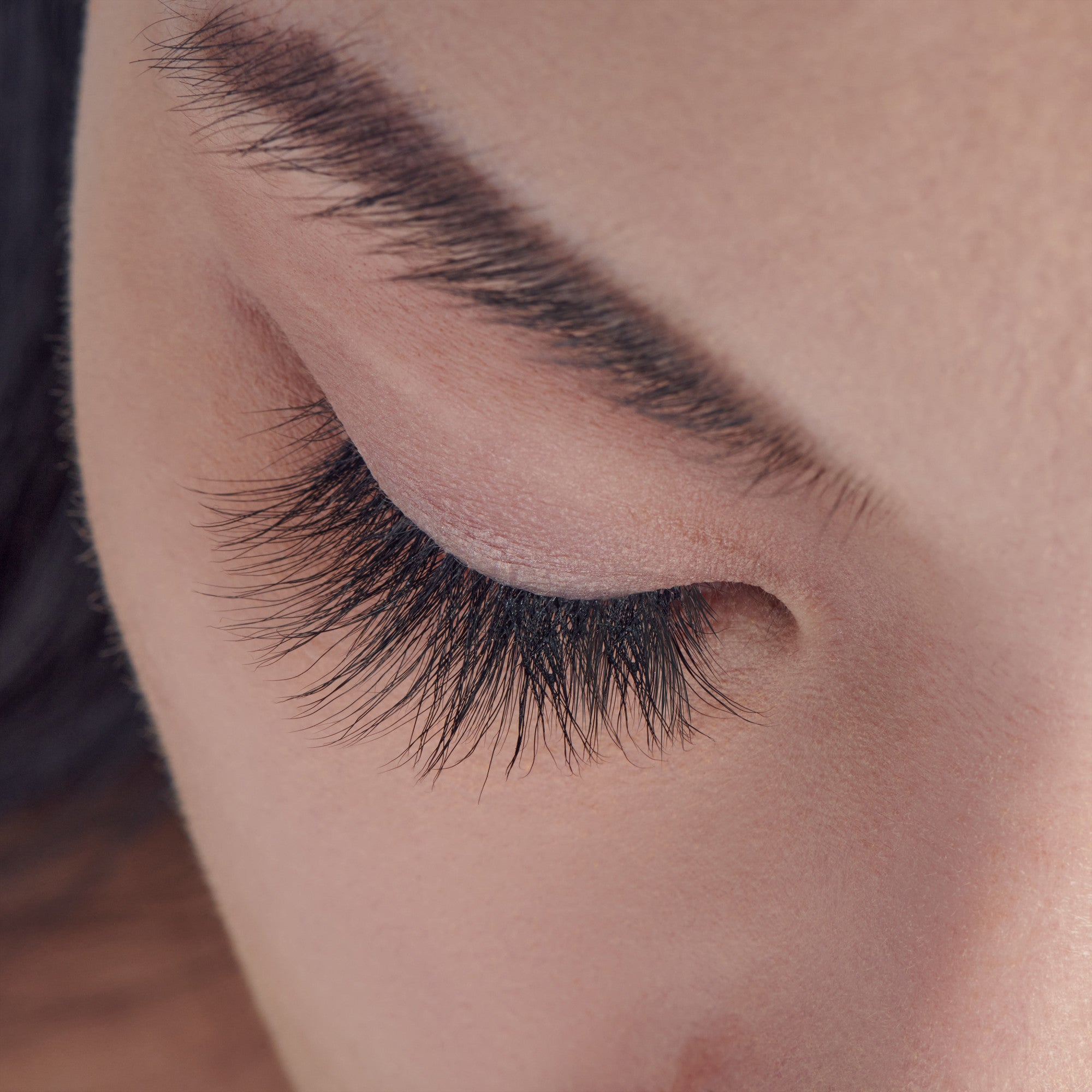 Close-up of a closed eye showing long, thick, dark, faux mink eyelash extensions enhancing natural lashes with a soft, natural skin tone background.