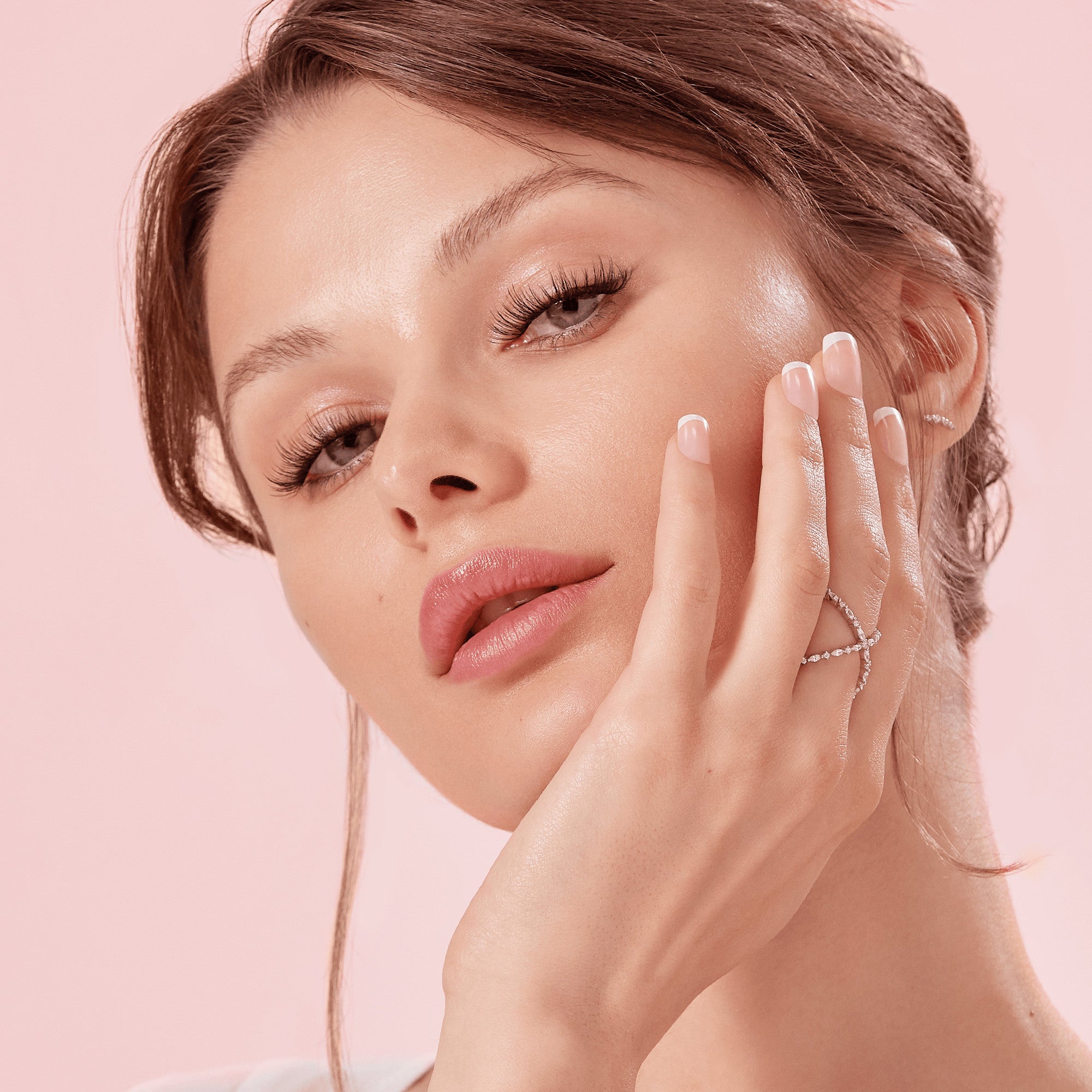 Close-up of a woman with light brown hair wearing long, voluminous self-adhesive false eyelashes and soft pink lip color, posing with her hand near her face against a light pink background.
