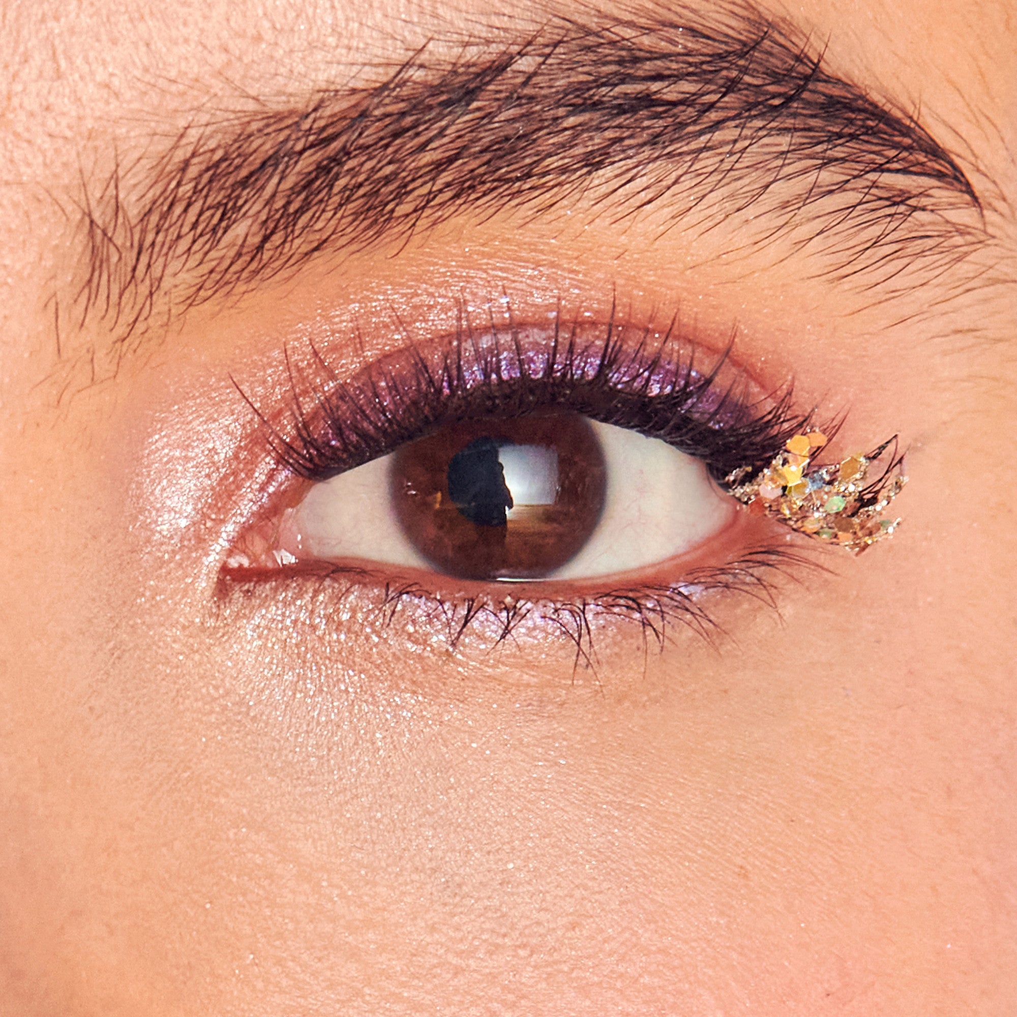 Close-up of a brown eye with shimmery pink and purple eyeshadow and a cluster of iridescent glitter flakes applied near the outer corner of the lower lashes.