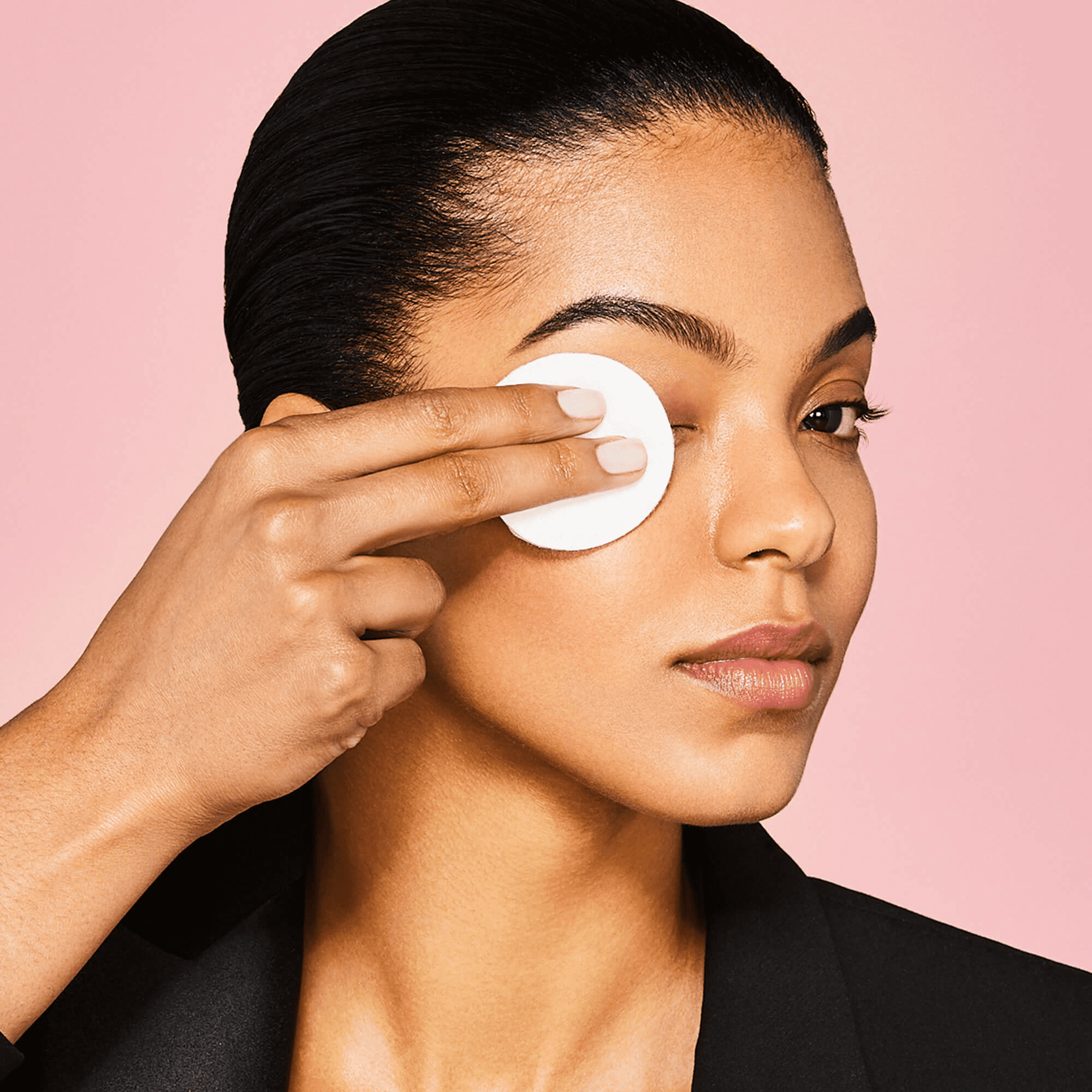 Woman with smooth skin and slicked-back hair gently wiping her closed eye with a white cotton pad against a pink background.
