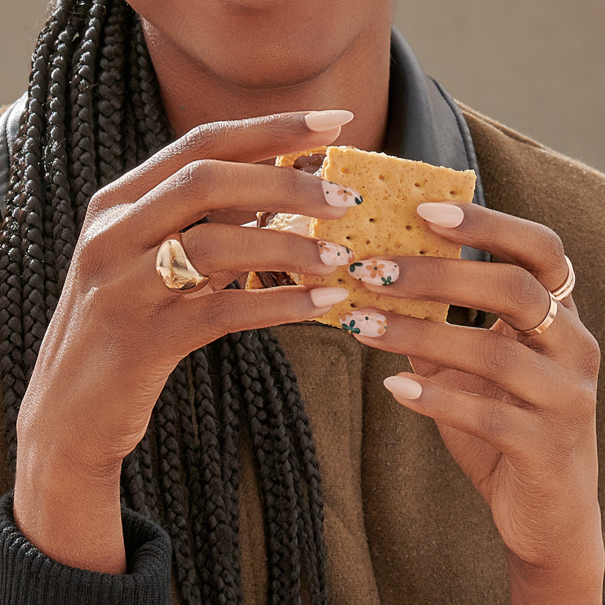 Hands with press-on nails holding a s’more. Nails are a mix of solid pale pink and floral designs with orange and dark green flowers on a sheer base.