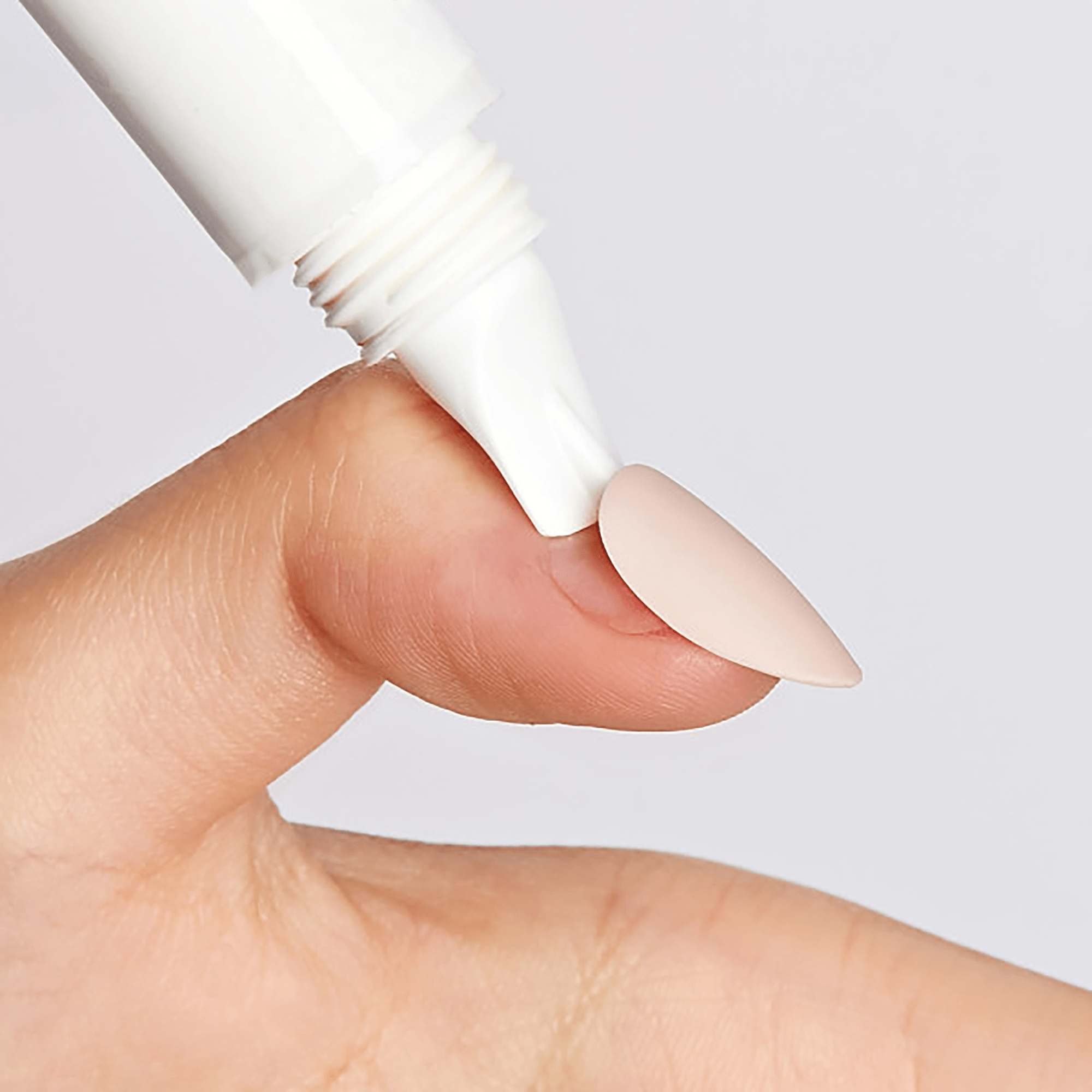 Fingertip with light pink almond-shaped press-on nail, with the tip of a white tube applicator positioned near the nail for glue removal.