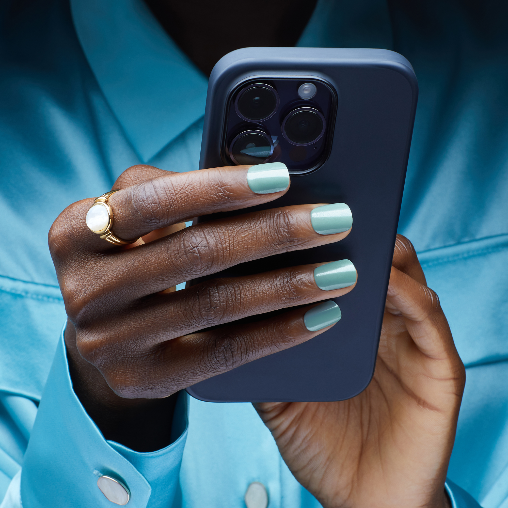 Hand with dark skin wearing a gold ring and light teal press-on nails holding a dark blue smartphone, against a blue shirt background.
