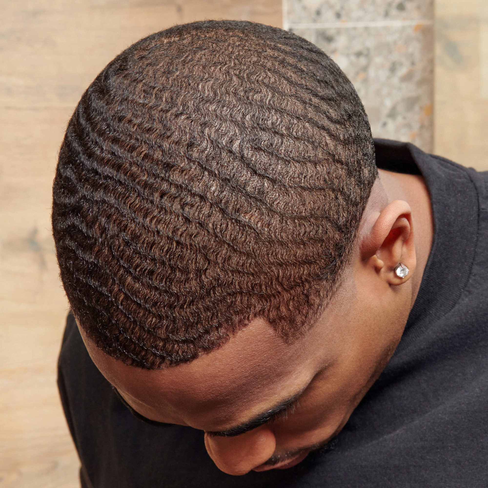 Close-up view of a man with well-defined 360 waves hairstyle, wearing a black shirt, head tilted downward against a neutral background.