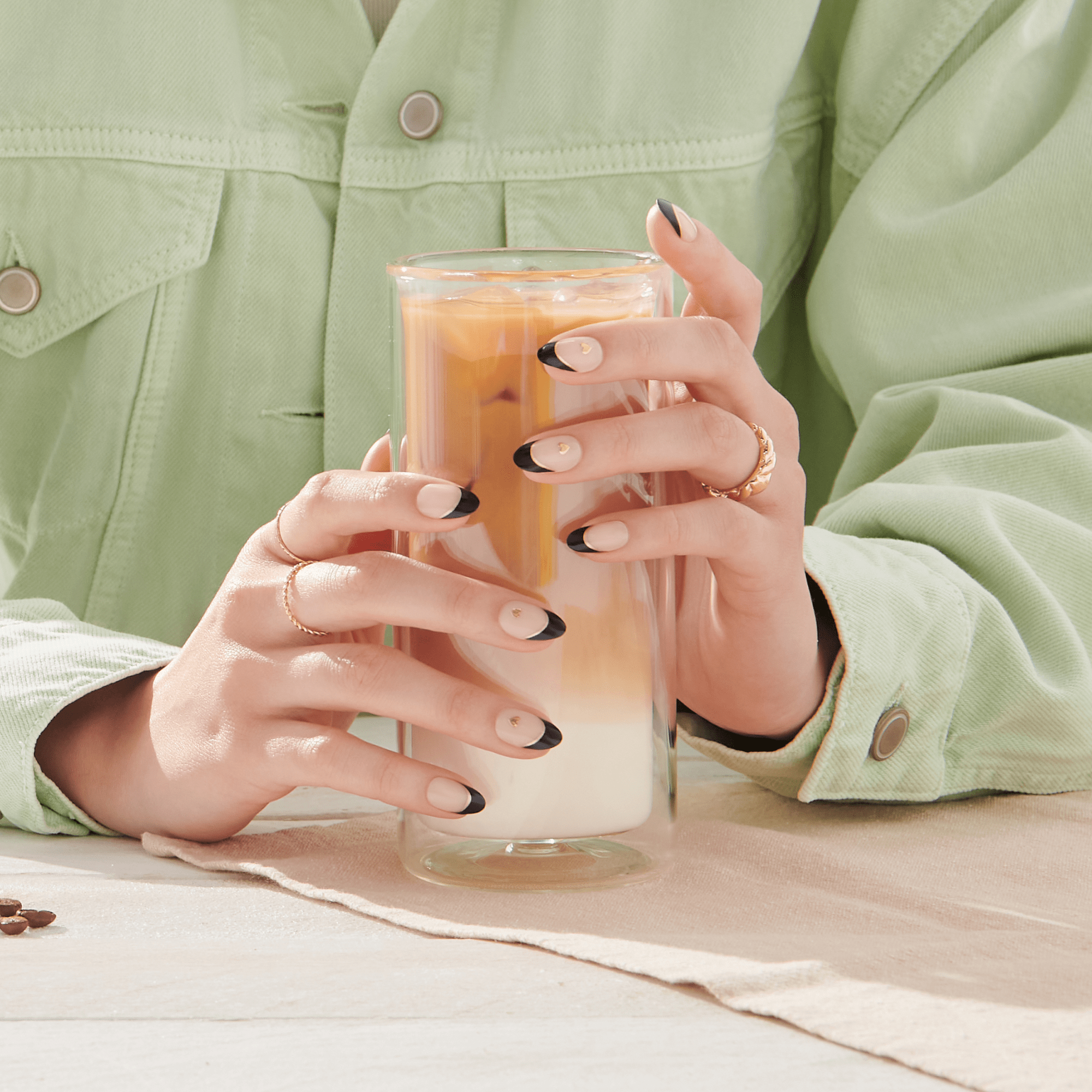 Hands with French latte press-on nails featuring nude base, black tips, and small gold heart accents hold a layered iced latte in a clear glass.