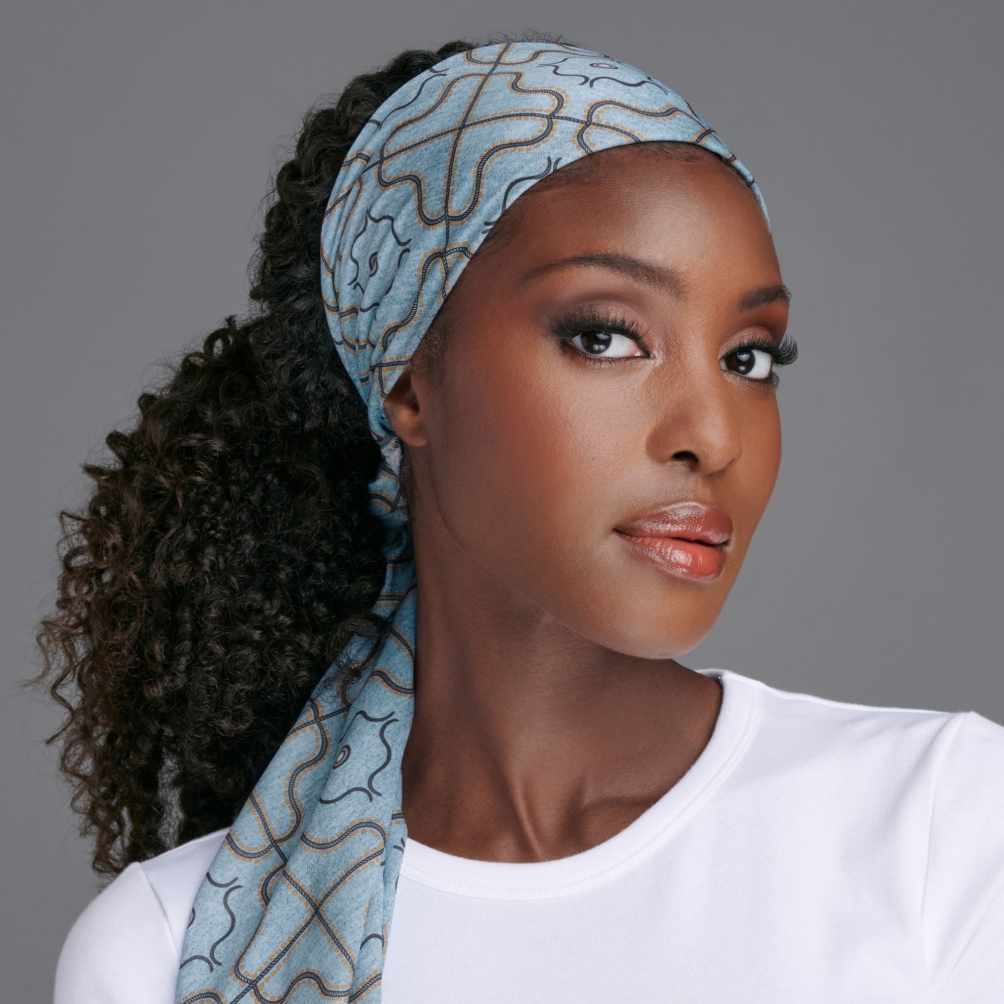 Close-up of a woman wearing a light blue satin wrap scarf with an abstract brown and black line pattern, styled over curly hair, paired with a white shirt.