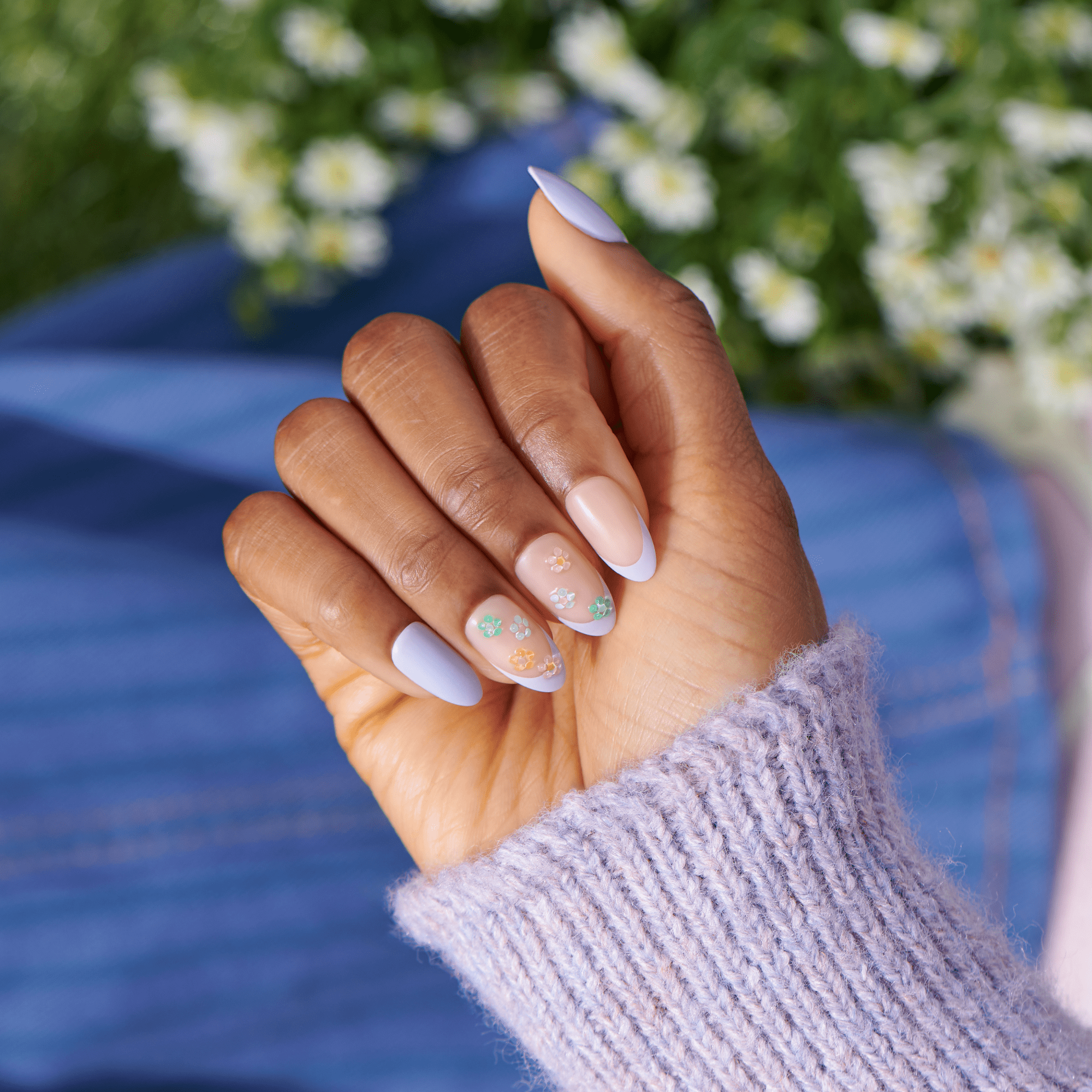 Lavender sweater hand displaying almond nails with white tips and floral nail art design outdoors.