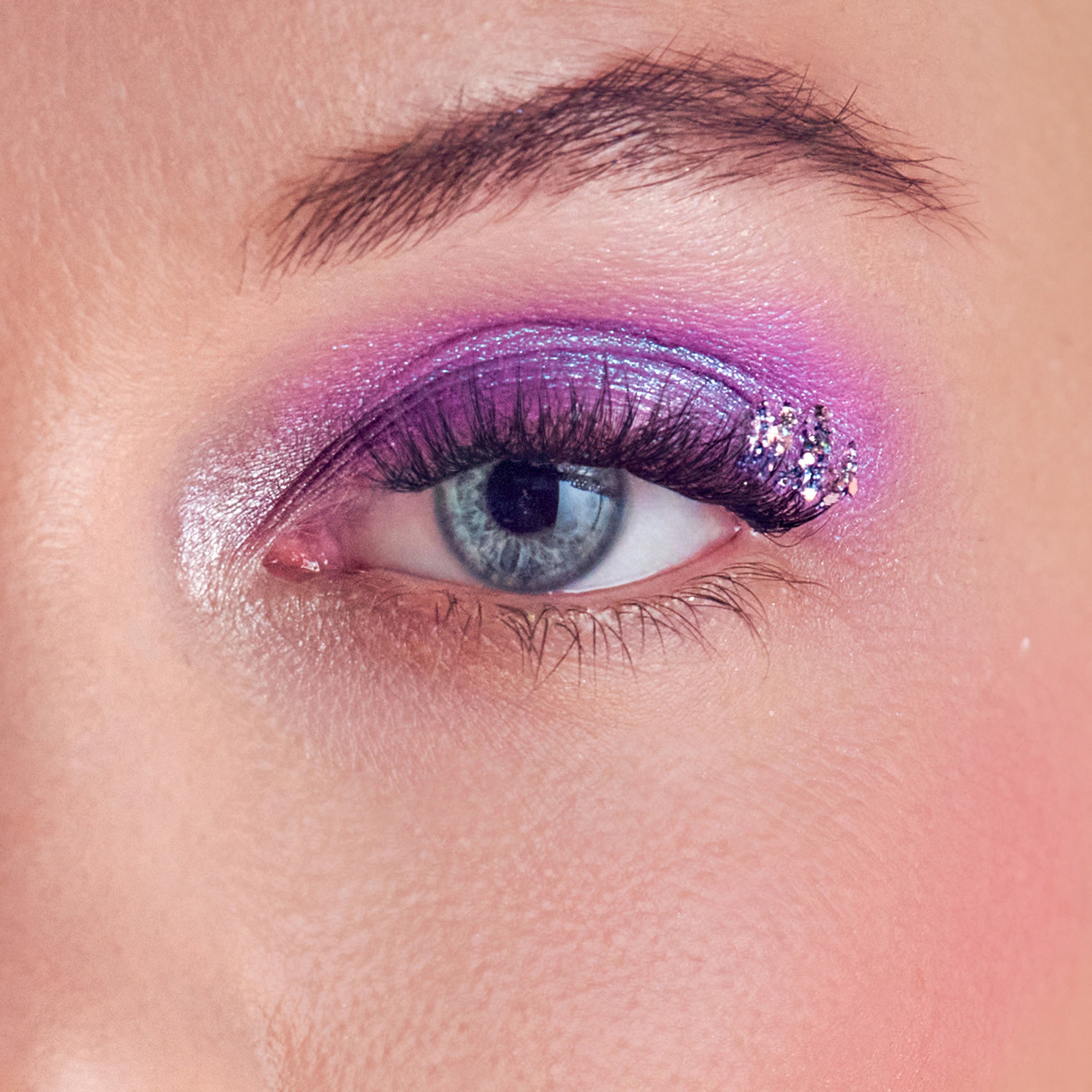 Close-up of a blue eye wearing long, dark false eyelashes with purple shimmery eyeshadow and glitter accents on the outer corner.