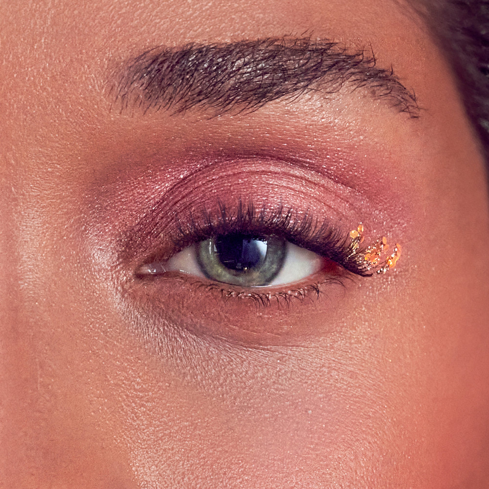 Close-up of a green eye with pink shimmery eyeshadow and decorative gold glitter accents on the outer corner of natural-looking false eyelashes.