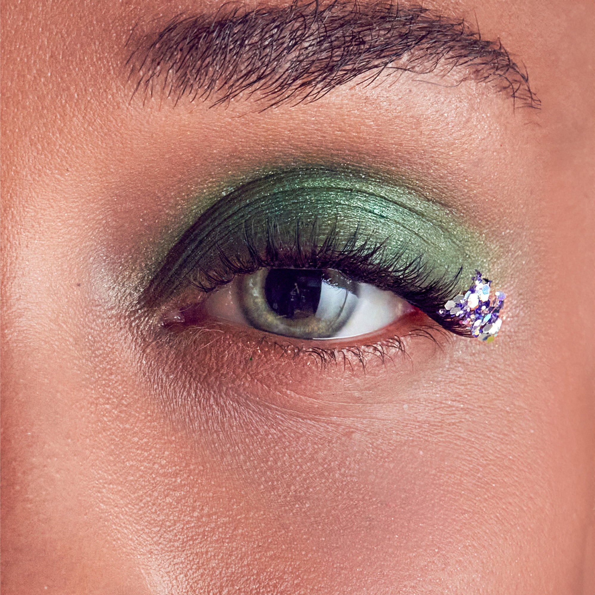 Close-up of an eye with light green metallic eyeshadow and long false eyelashes featuring a decorative cluster of purple and silver glitter at the outer corner.
