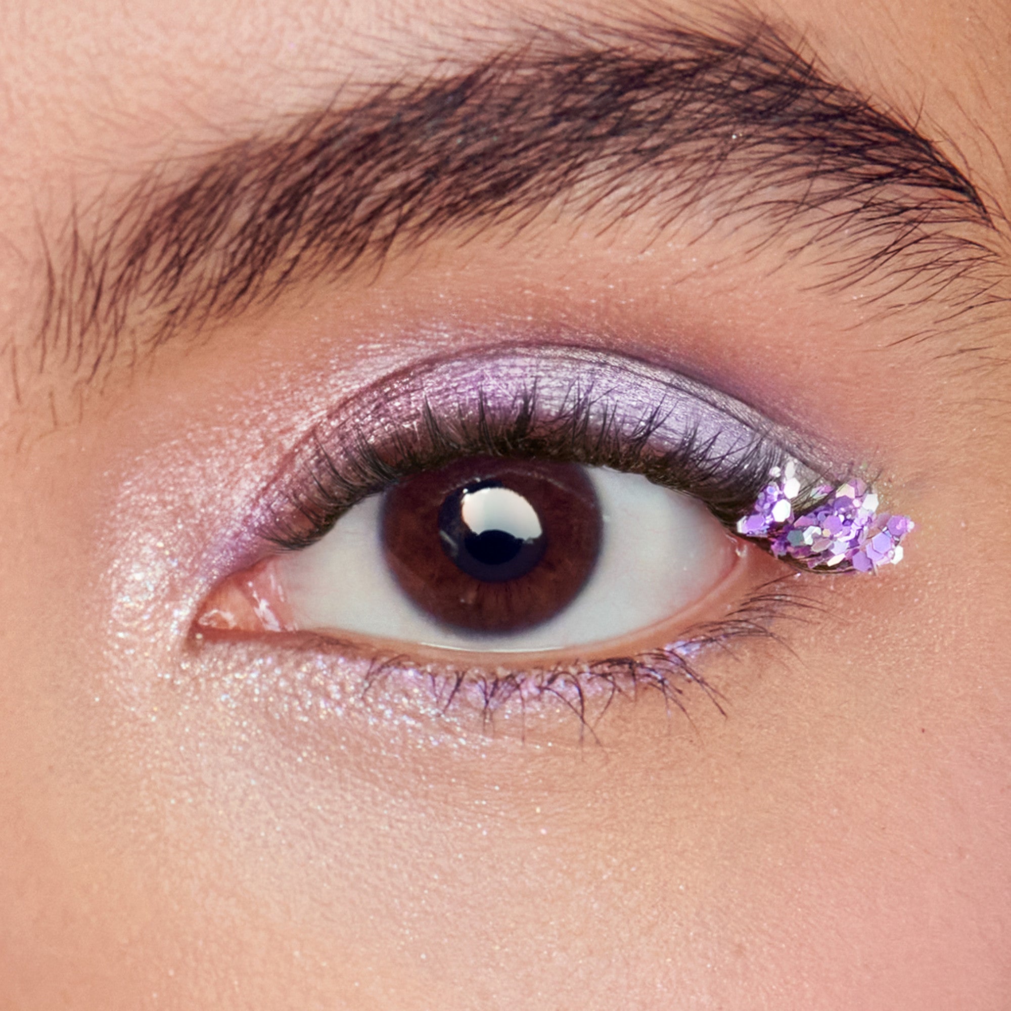 Close-up of a brown eye with shimmery lavender eyeshadow and small purple metallic hexagon confetti applied at the outer corner on the upper eyelid.