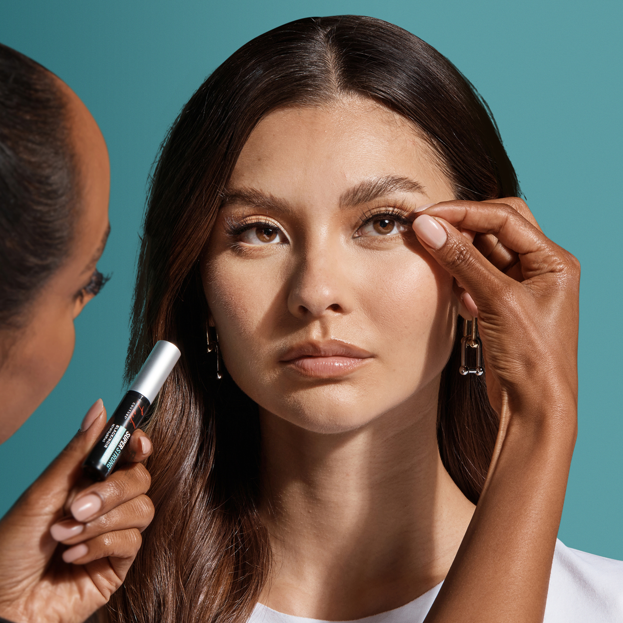 A person with medium skin tone applies a black tube of KISS Lash Couture Strip Lash Adhesive to the upper lash line of a woman with long dark brown hair and neutral makeup against a teal background.
