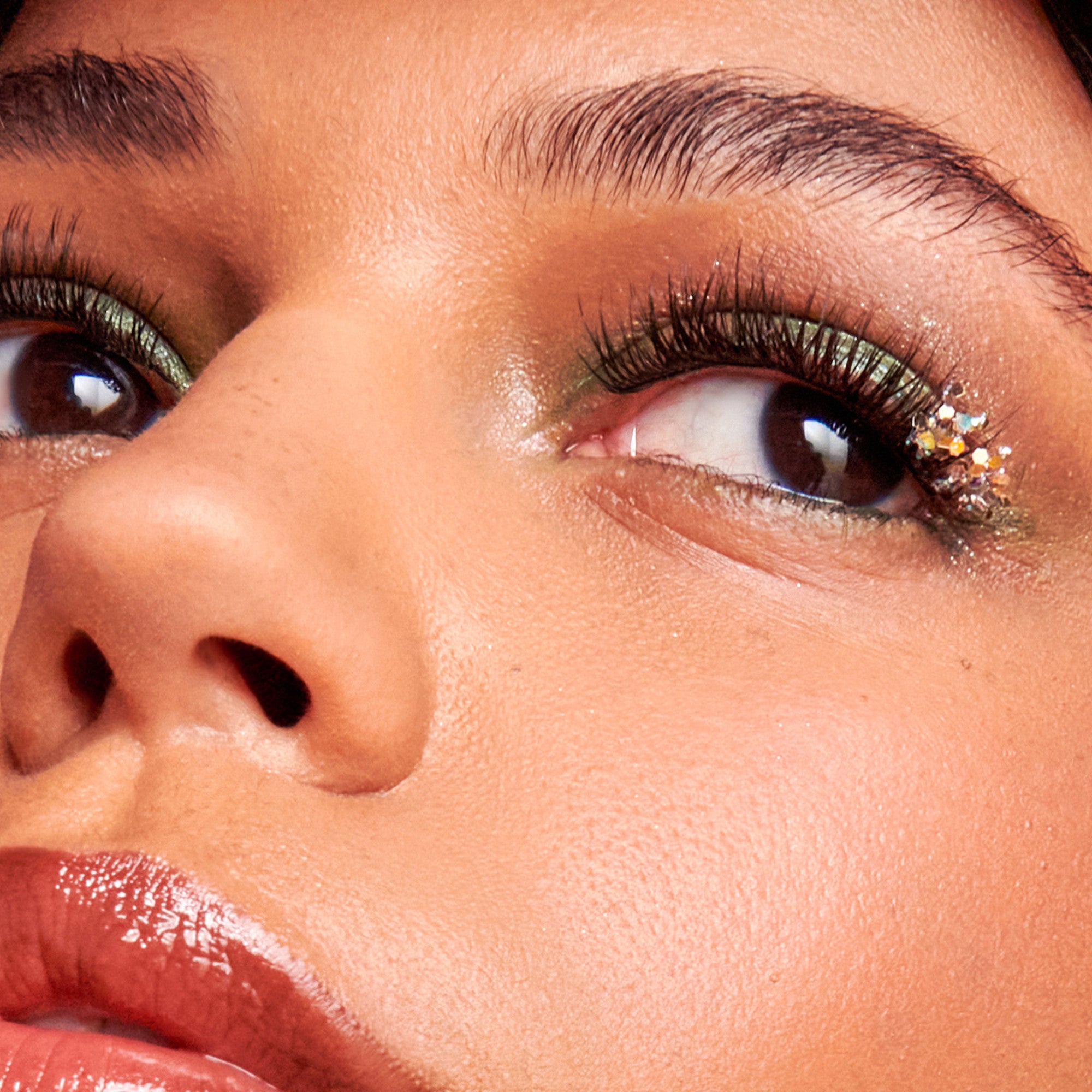 Close-up of a person’s eye with green metallic eyeshadow, long lashes, and small iridescent rhinestones clustered at the outer corner, glossy lips visible.