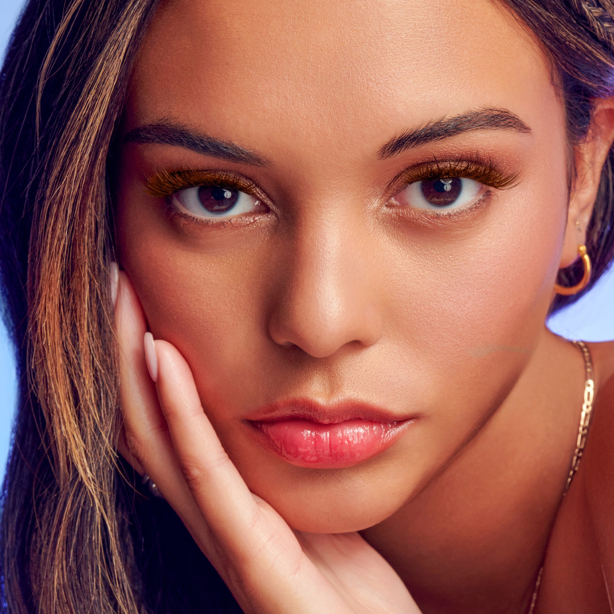 Close-up of a woman with warm brown false eyelashes accentuating her eyes, natural makeup, glossy lips, and light brown highlighted hair against a soft purple background.
