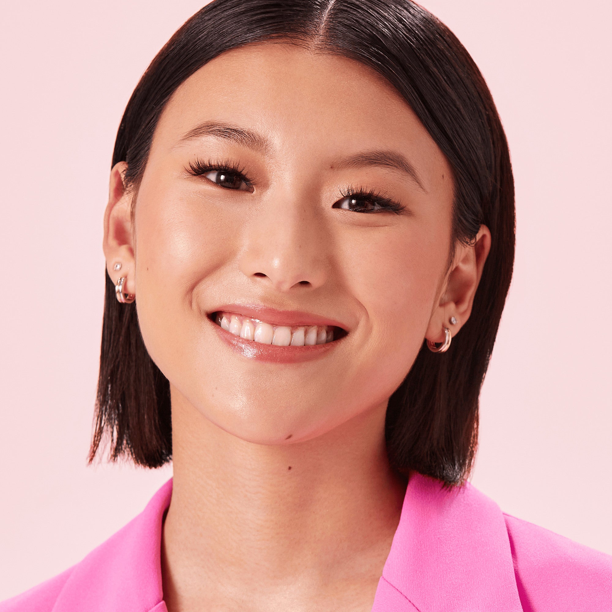 Close-up of a smiling woman with straight dark hair wearing natural-looking, voluminous false eyelashes and a bright pink blazer against a light pink background.