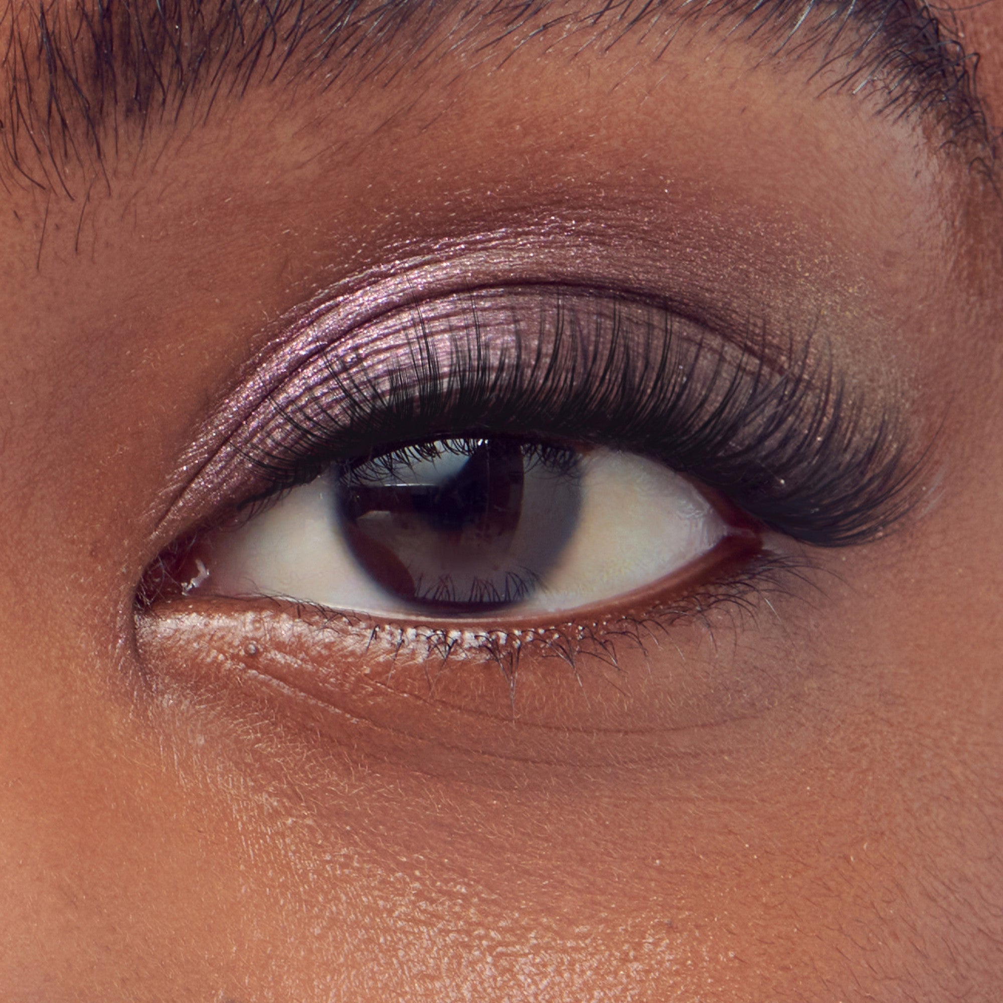 Close-up of an eye with long, wispy self-adhesive false eyelashes applied, featuring soft shimmery pink eyeshadow on the eyelid.