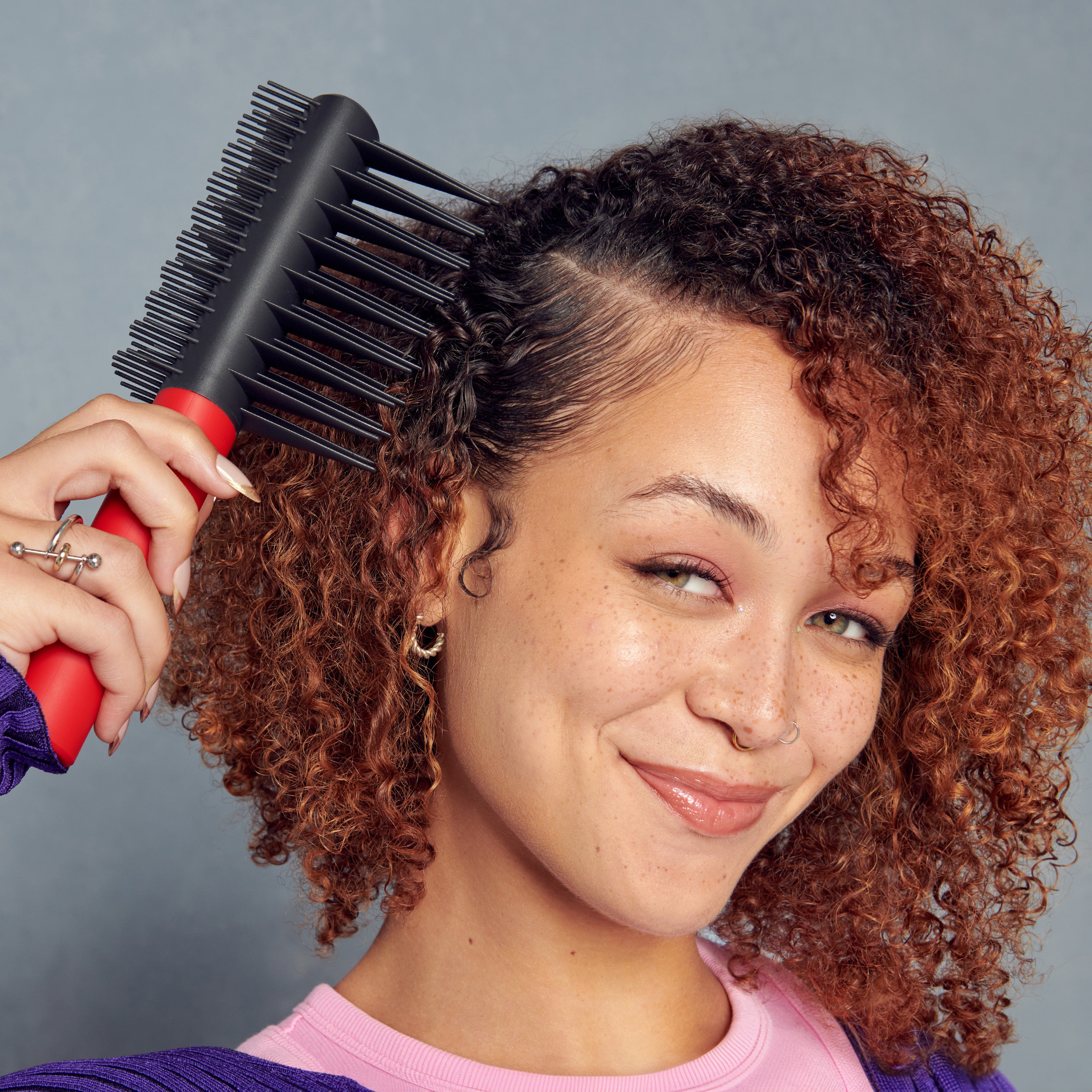 Young woman with curly reddish-brown hair holding a red and black detangling brush near her head, smiling against a gray background.